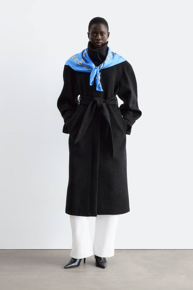 A long black belted coat with structured shoulders and a high collar is being worn by the model. The model is standing with their hands in the coat pockets, styling the look with a blue patterned scarf tied around the collar, white trousers, and black boots. - 1