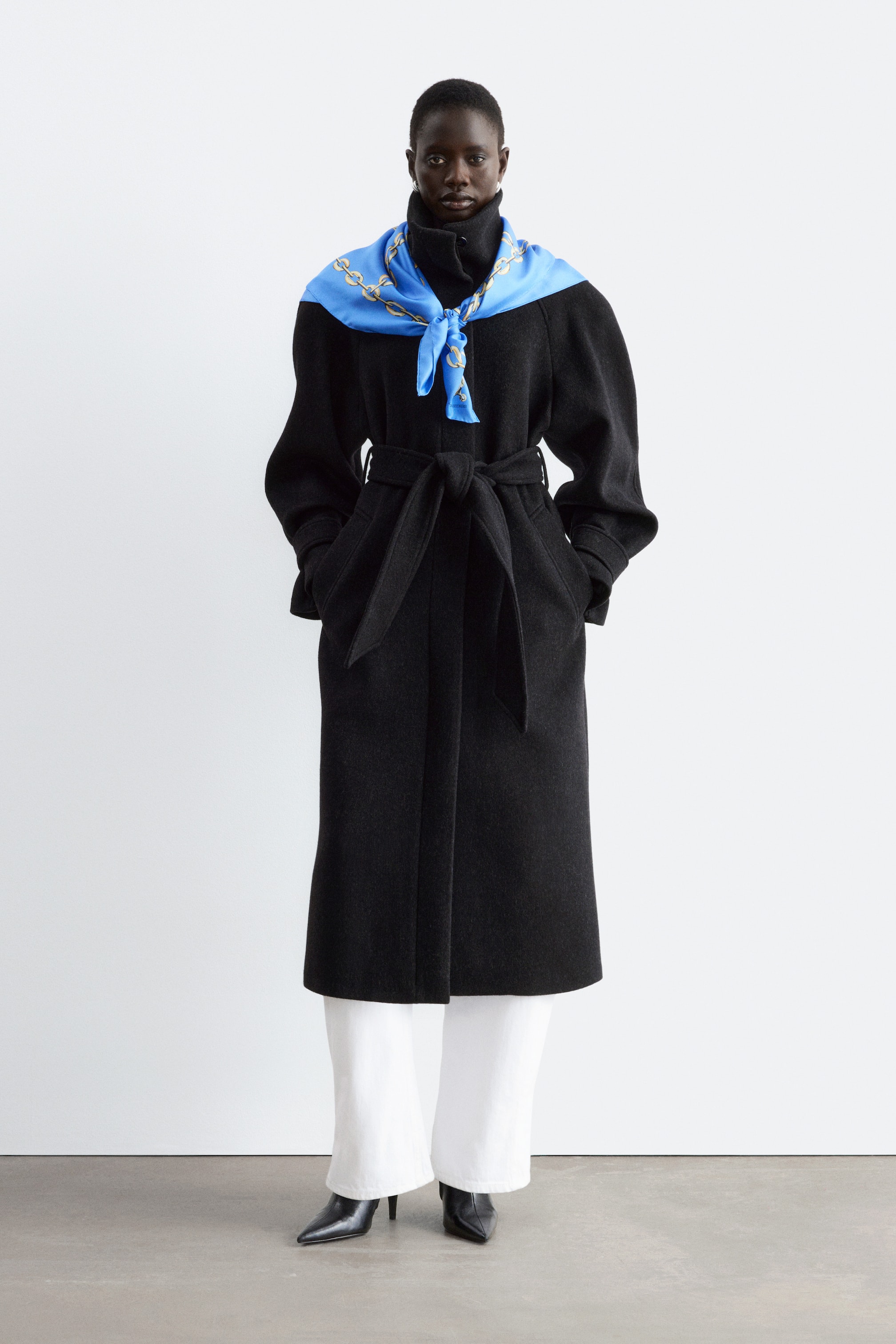 Größeres Bild ansehen: Ein langer, schwarzer Mantel mit Gürtel, strukturierten Schultern und hohem Kragen wird vom Model getragen. Das Model steht mit den Händen in den Taschen des Mantels und stylt den Look mit einem blau gemusterten Schal, der um den Kragen gebunden ist, weißen Hosen und schwarzen Stiefeln.
