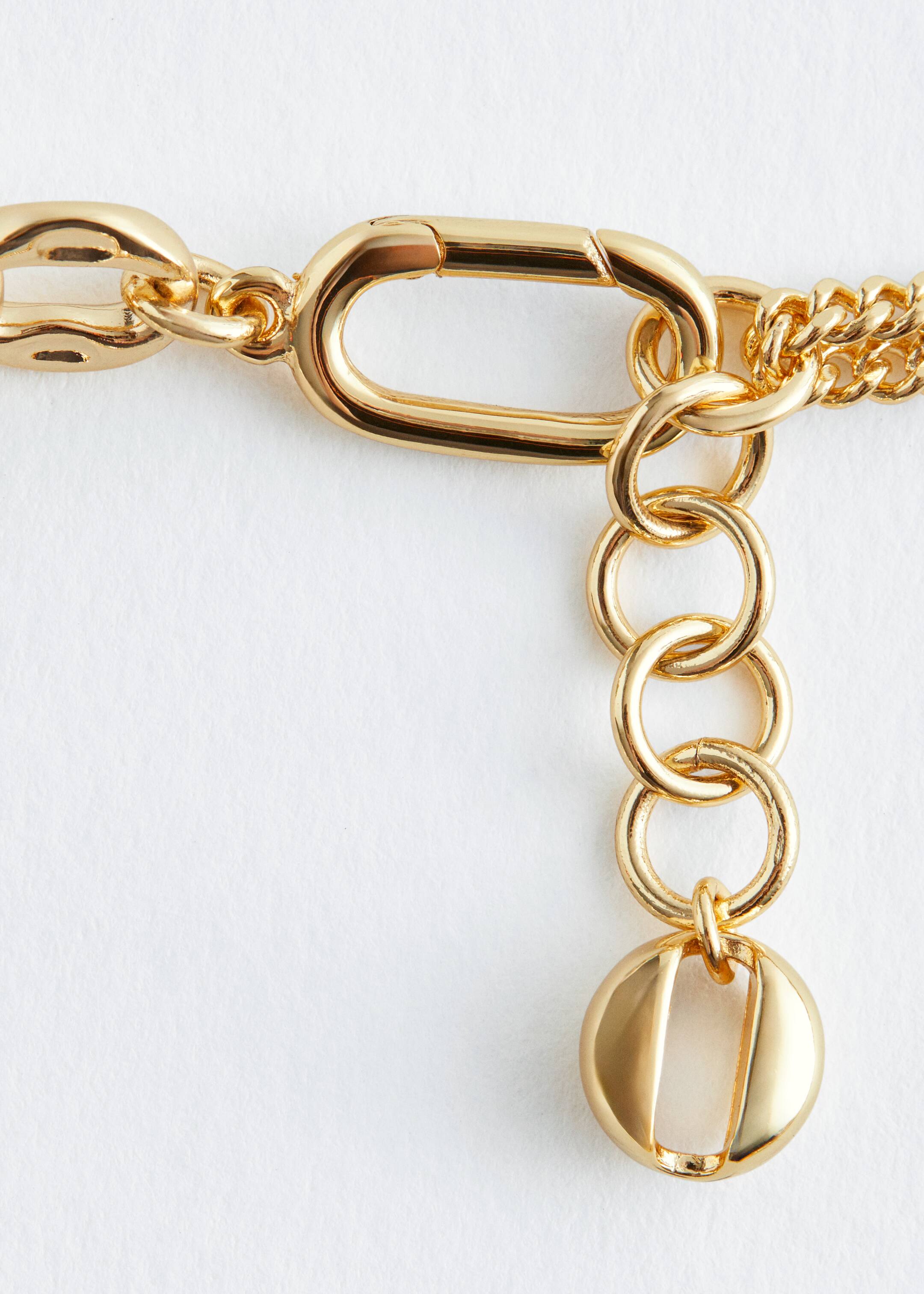 View larger image: A gold-colored necklace is presenting a close-up of its elongated, carabiner-style clasp that is connecting to an extension chain. The extension chain is made up of four round links, ending with a flat, round charm that is featuring a central vertical cut-out, while the main necklace chain, made of interlocking elongated links, is visible on the left.