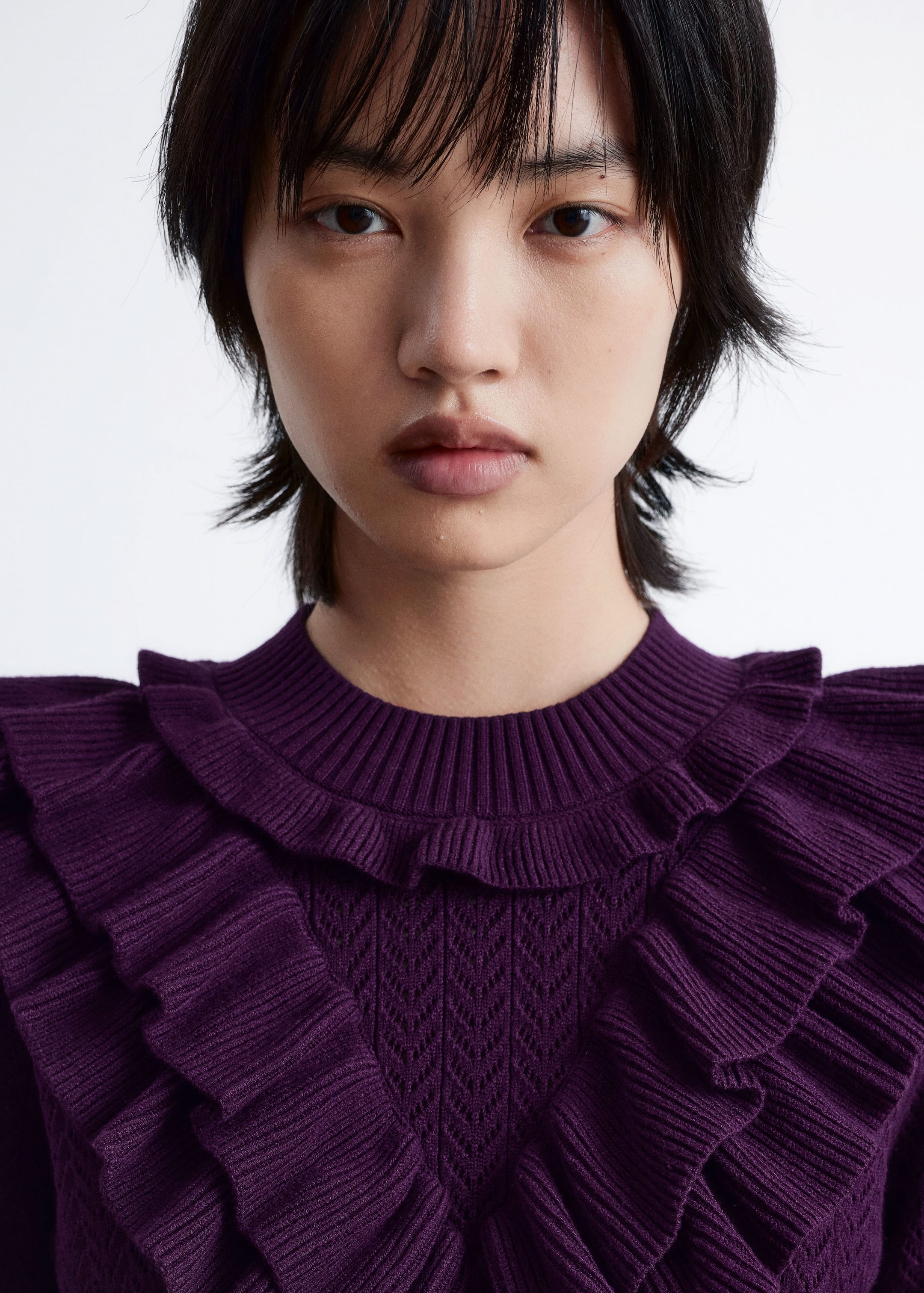 View larger image: A deep purple knit sweater is being displayed in a close-up, front view, featuring a ribbed crew neck and an openwork knit pattern on the main body. Prominent, cascading ribbed ruffles are extending across the neckline, chest, and shoulders.