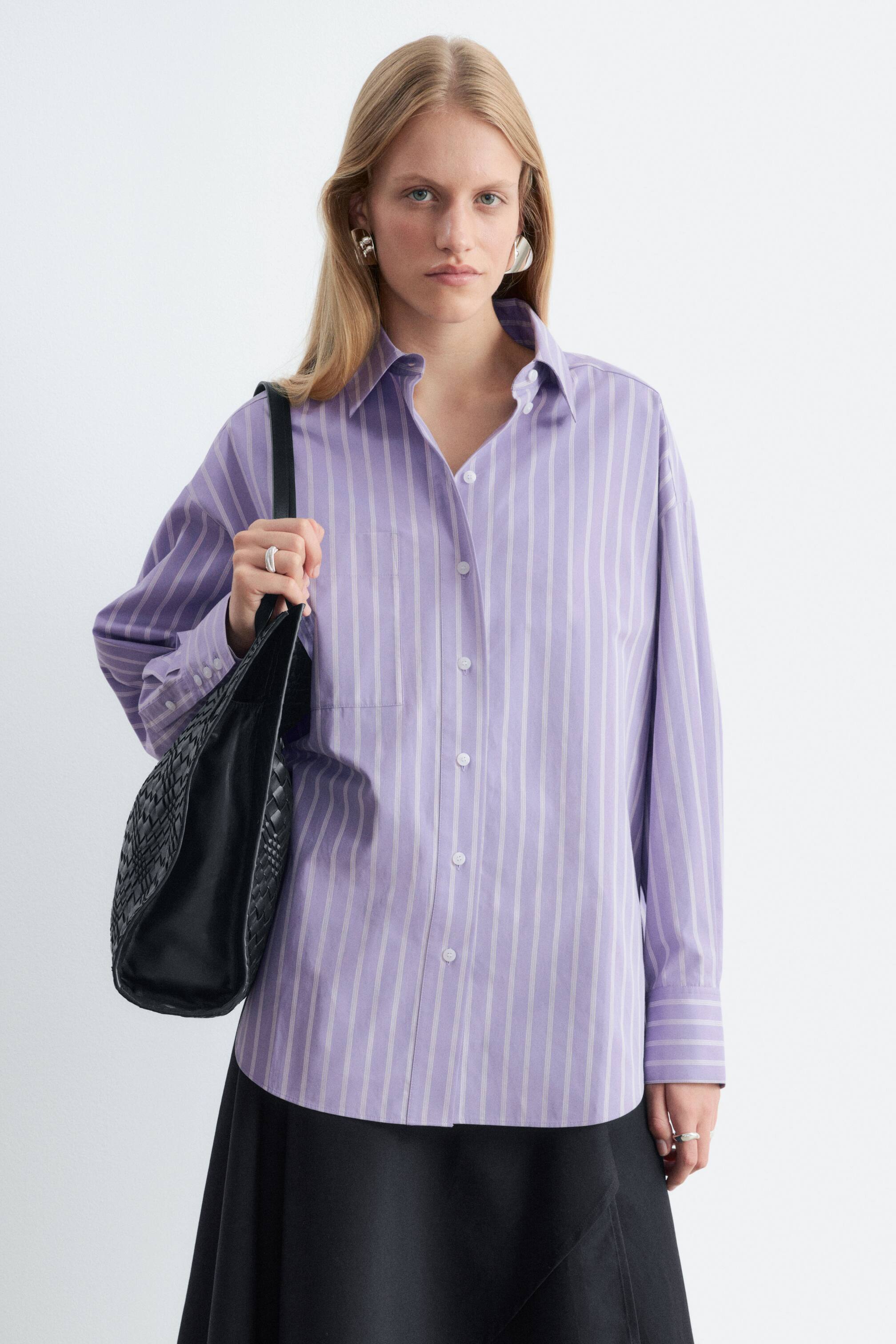 Näytä suurempi kuva: Mallilla on yllään oversized-tyylinen, vaaleanvioletti-valkoraidallinen pitkähihainen paita, jossa on terävä kaulus, napitus edessä ja rintatasku. Paita on stailattu mustan hameen kanssa, ja mallilla on musta punottu olkalaukku.