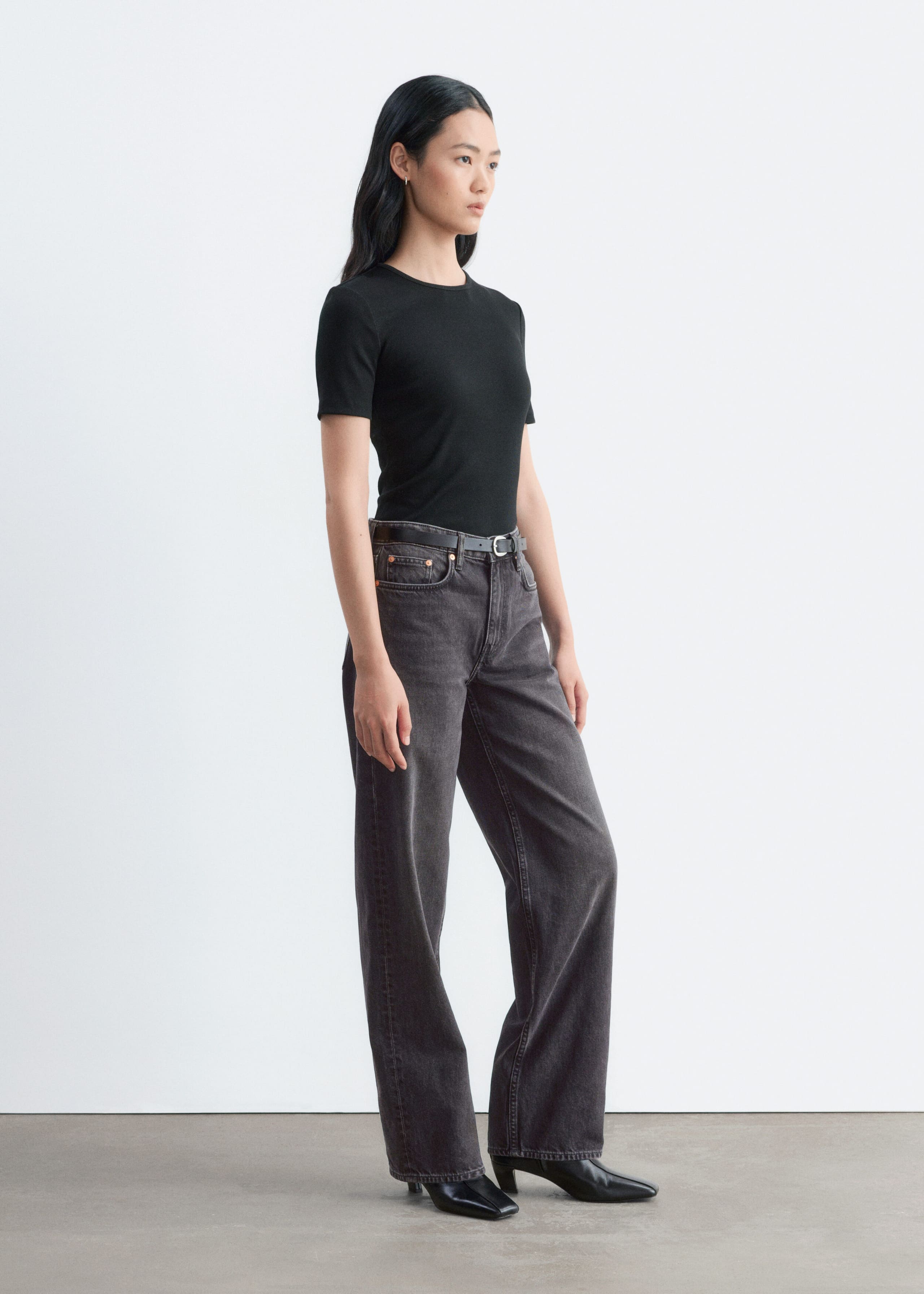 A black, ribbed slim-fit T-shirt featuring a crew neck and short sleeves is being worn by the model, tucked into dark grey wide-leg jeans.