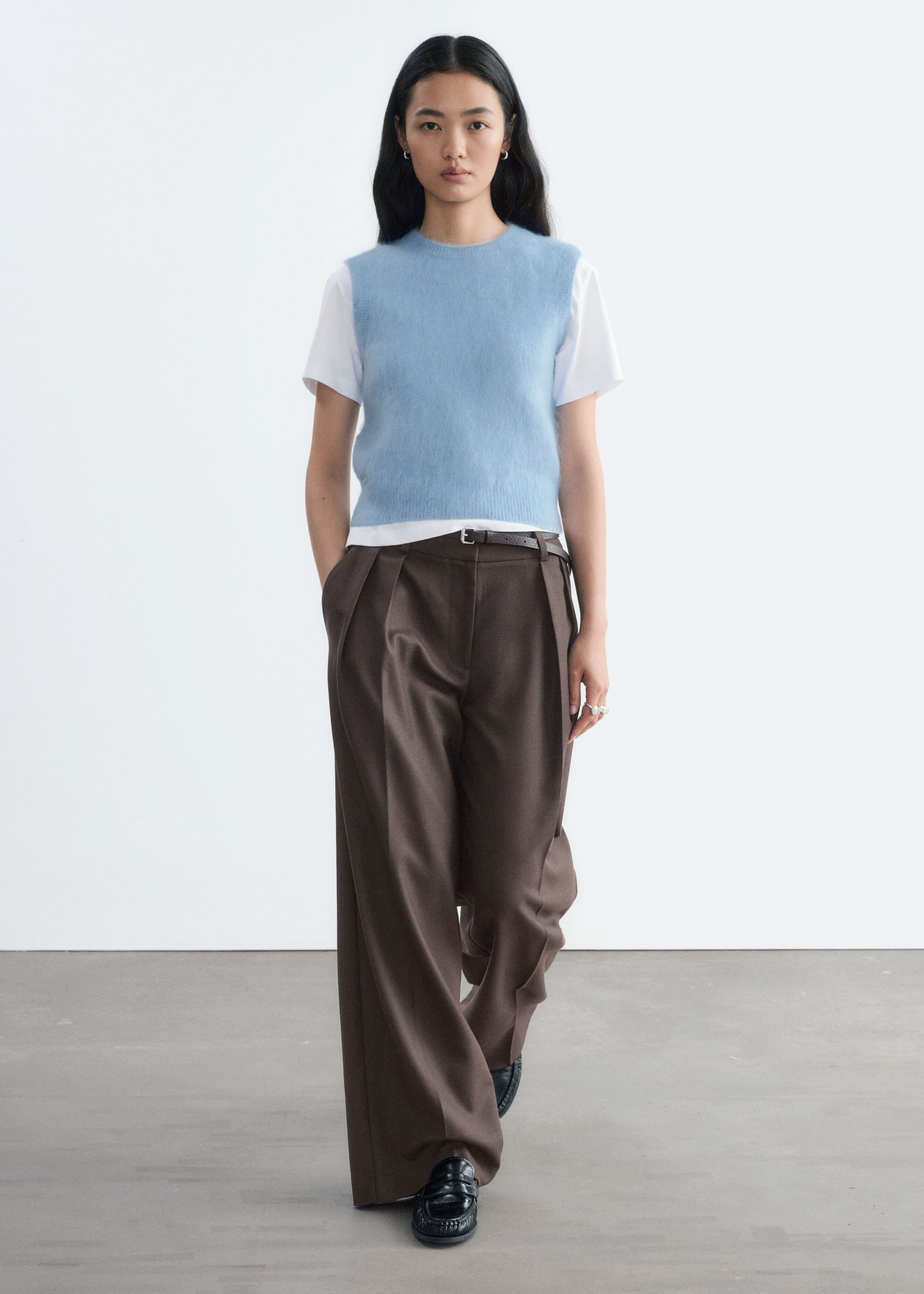 Un chaleco de mezcla de mohair de punto en azul claro, con cuello redondo y una textura suave y esponjosa, lo lleva la modelo. El chaleco sin mangas se lleva sobre una camiseta blanca de manga corta y se combina con pantalones marrones de pierna ancha.