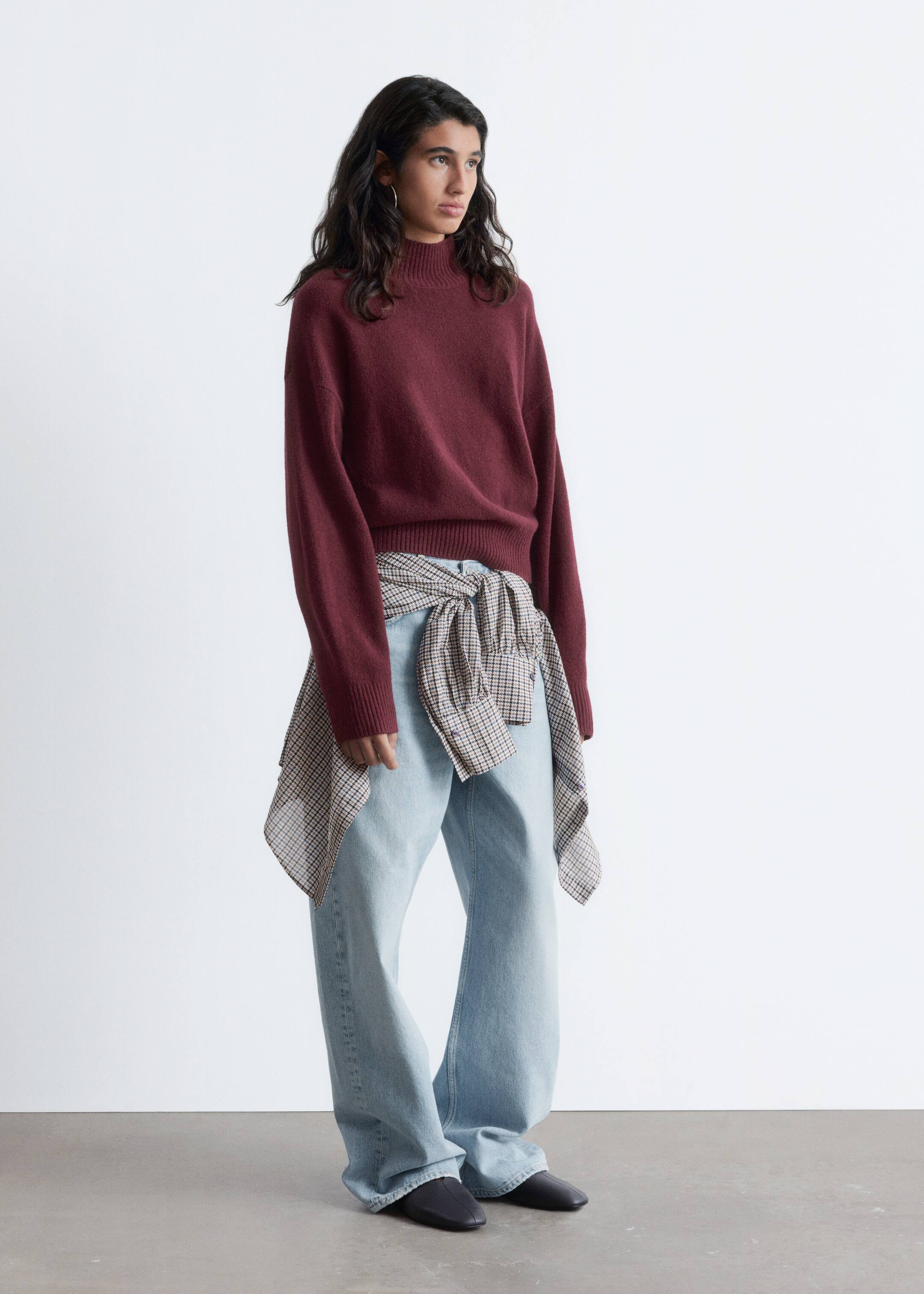 View larger image: A burgundy mock-neck sweater is being displayed from a slight right angle, showing its relaxed fit with visible ribbed cuffs and a ribbed hem. It is being styled with light wash, wide-leg jeans and a beige and brown checkered shirt tied around the waist.