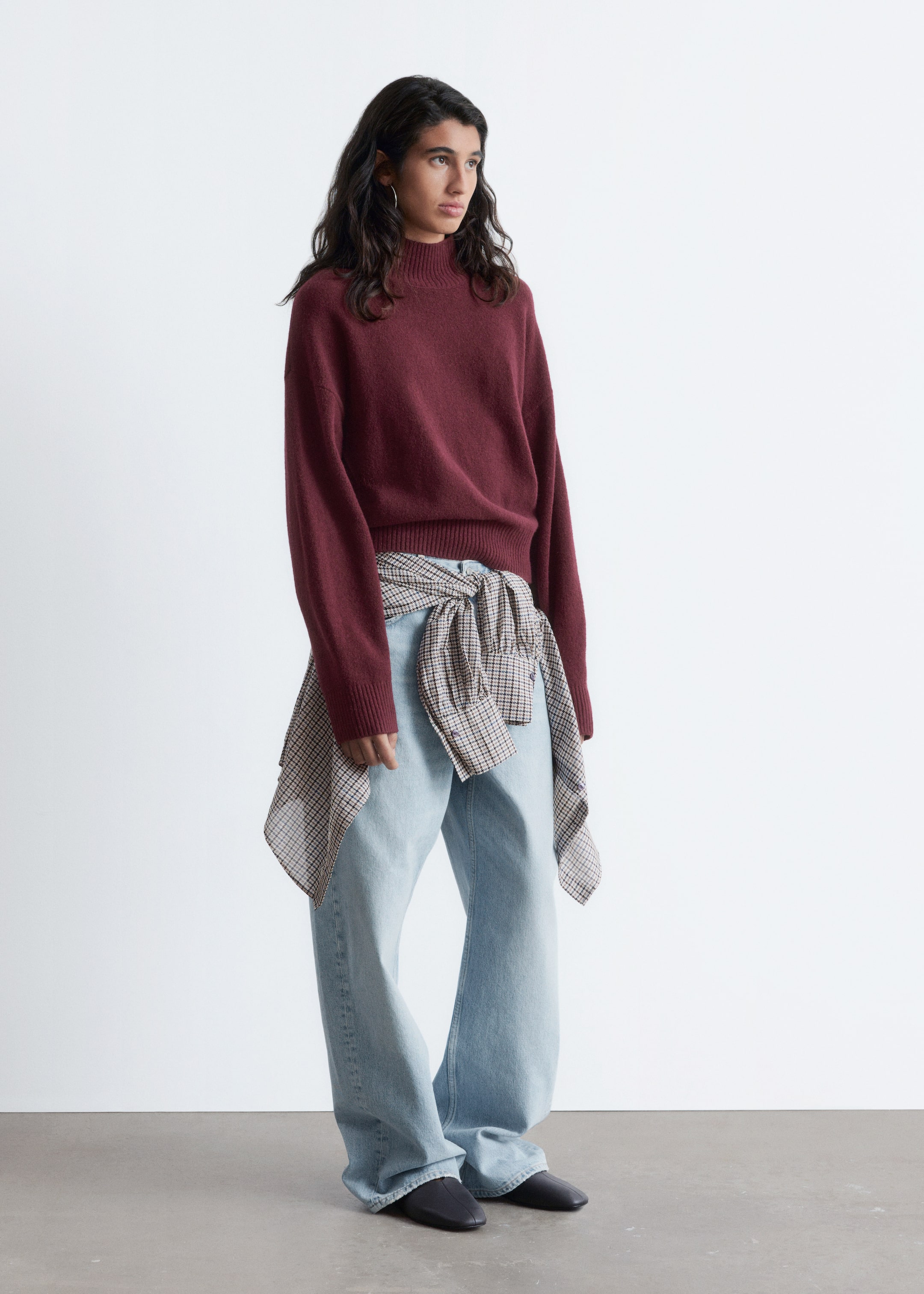 Größeres Bild anzeigen: Ein bordeauxroter Mock-Neck-Pullover wird aus einem leichten rechten Winkel gezeigt, der seine entspannte Passform mit sichtbaren gerippten Bündchen und einem gerippten Saum zeigt. Er wird mit hell gewaschenen, weit geschnittenen Jeans und einem beige-braunen karierten Hemd, das um die Taille gebunden ist, gestylt.