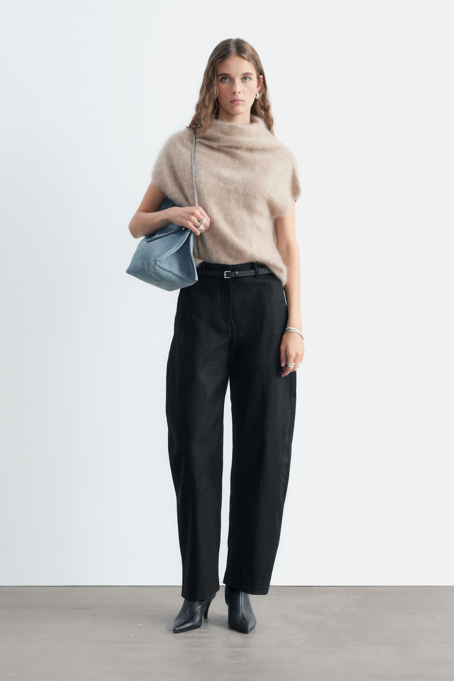 La modella indossa pantaloni barrel leg in twill di cotone nero, caratterizzati da vita alta e una silhouette ampia e strutturata con gamba curva. I pantaloni sono abbinati a una sottile cintura nera, un top in maglia senza maniche beige e stivali neri con tacco e punta affusolata. - 1