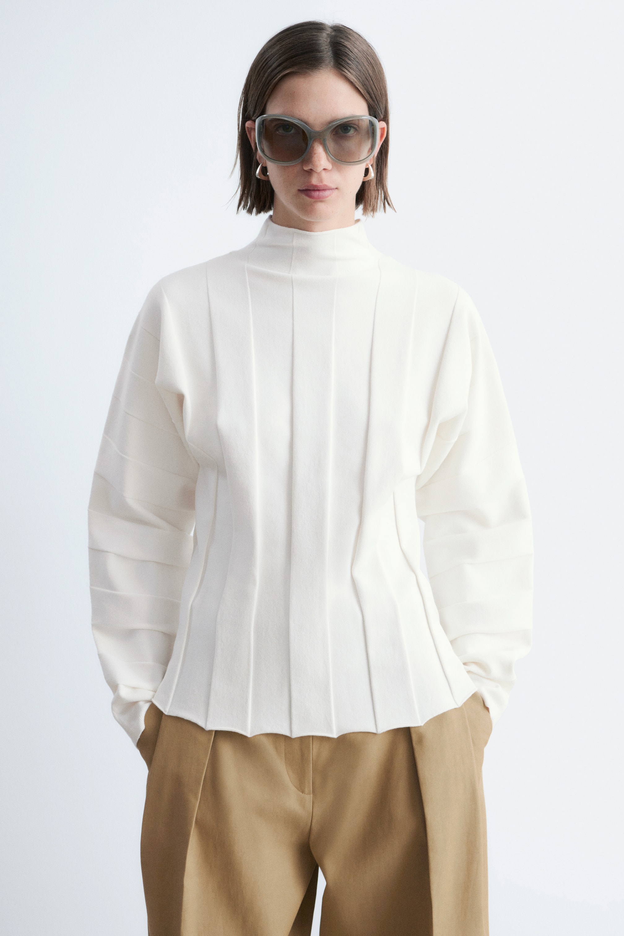 Zobacz większy obraz: Kremowy top z wysokim dekoltem i długimi rękawami z poziomymi zakładkami. Góra prezentuje wyraźne pionowe zakładki, które modelują talię i kończą się falistym brzegiem.