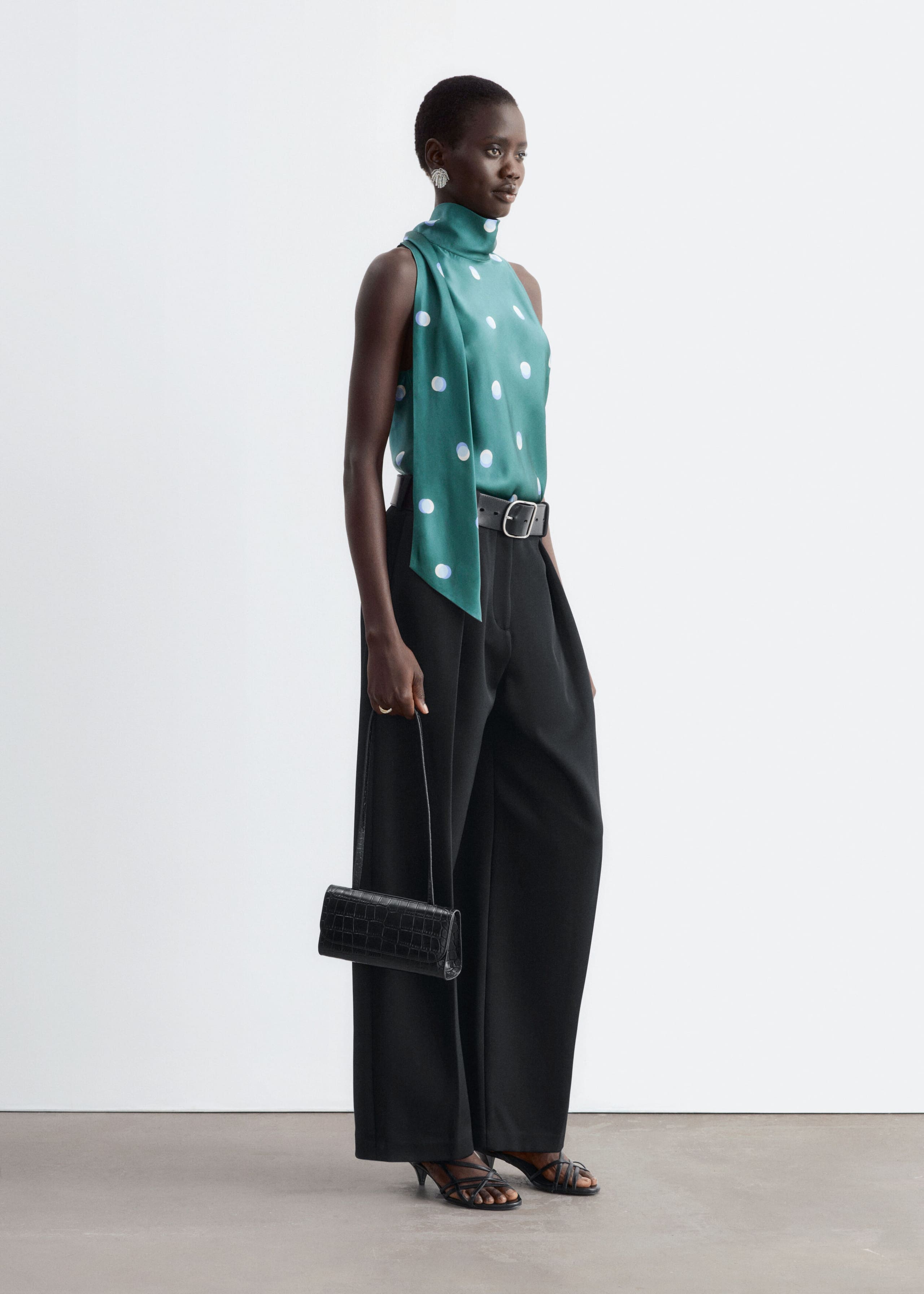 A green satin polka dot top with a high neck and long draped scarf detail is worn by the model. The sleeveless top is tucked into high-waisted, wide-leg black trousers secured with a wide belt.