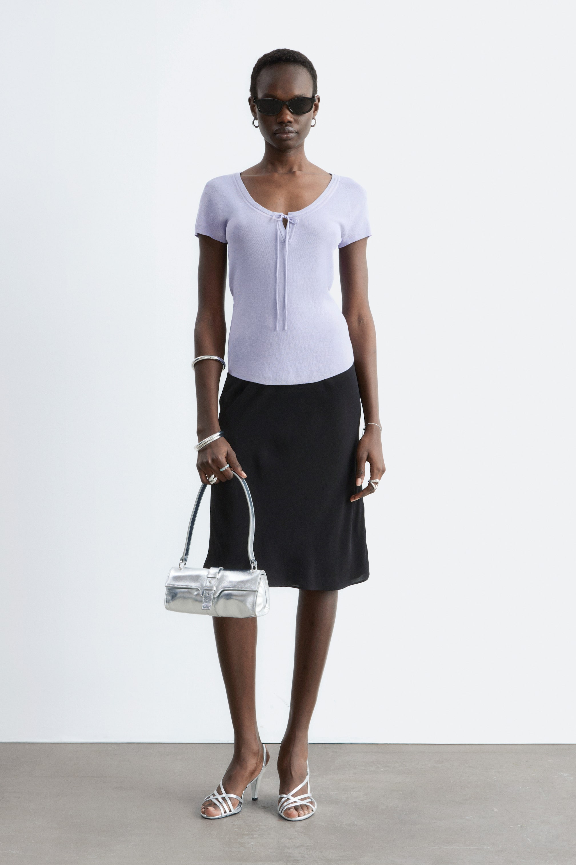 Ver imagem maior: A modelo está a usar uma saia preta de cetim. Apresenta uma silhueta reta com uma bainha ligeiramente evasé, caindo até ao comprimento do joelho.