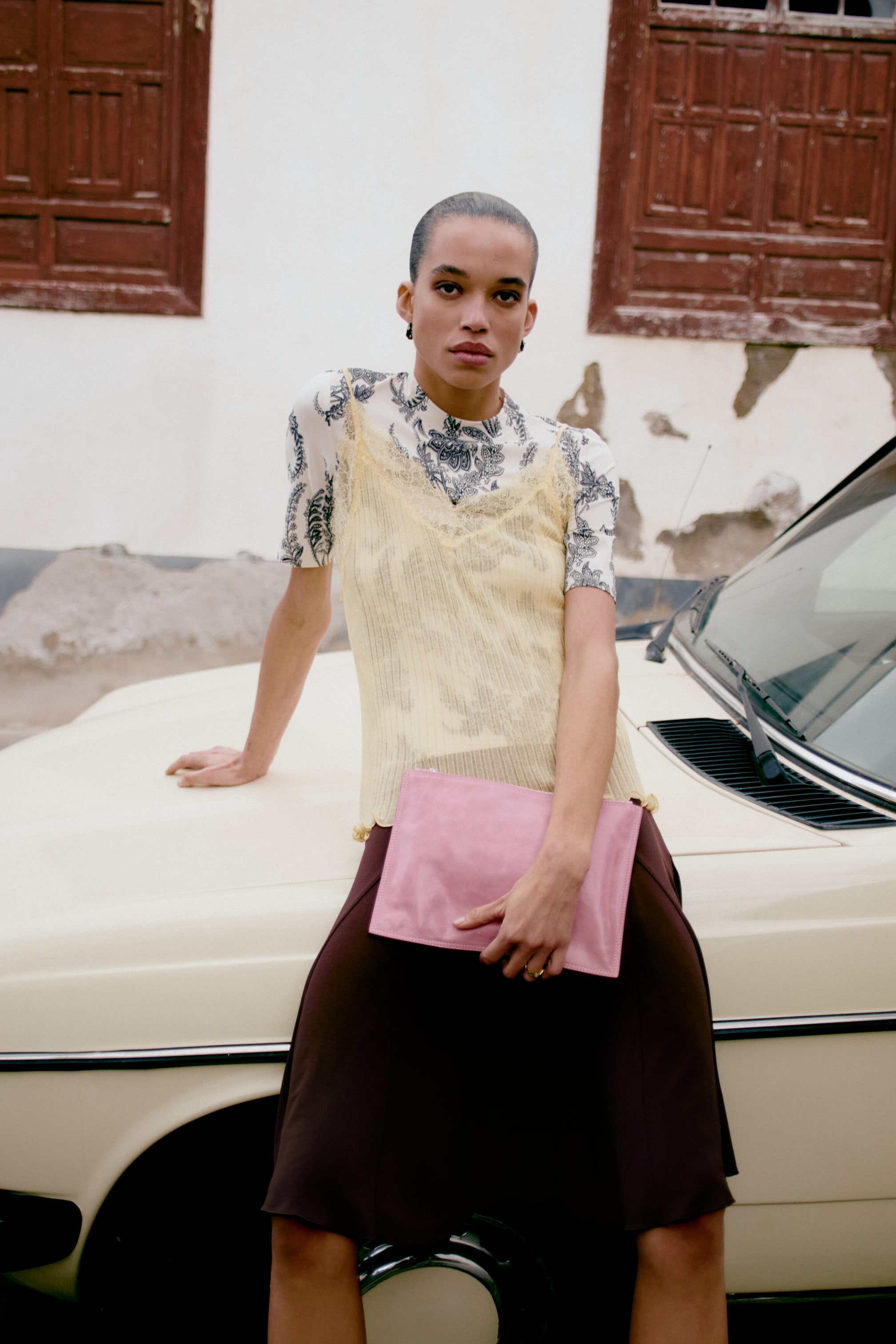 Se større billede: En hvid kortærmet top med sort-hvidt paisleymønster og høj halsudskæring bæres under en lysegul ribbet camisole, der har en V-udskæring med blondekant. En mørkebrun A-formet nederdel komplementerer de lagdelte toppe, og en rektangulær lyserød clutch taske holdes i venstre hånd.