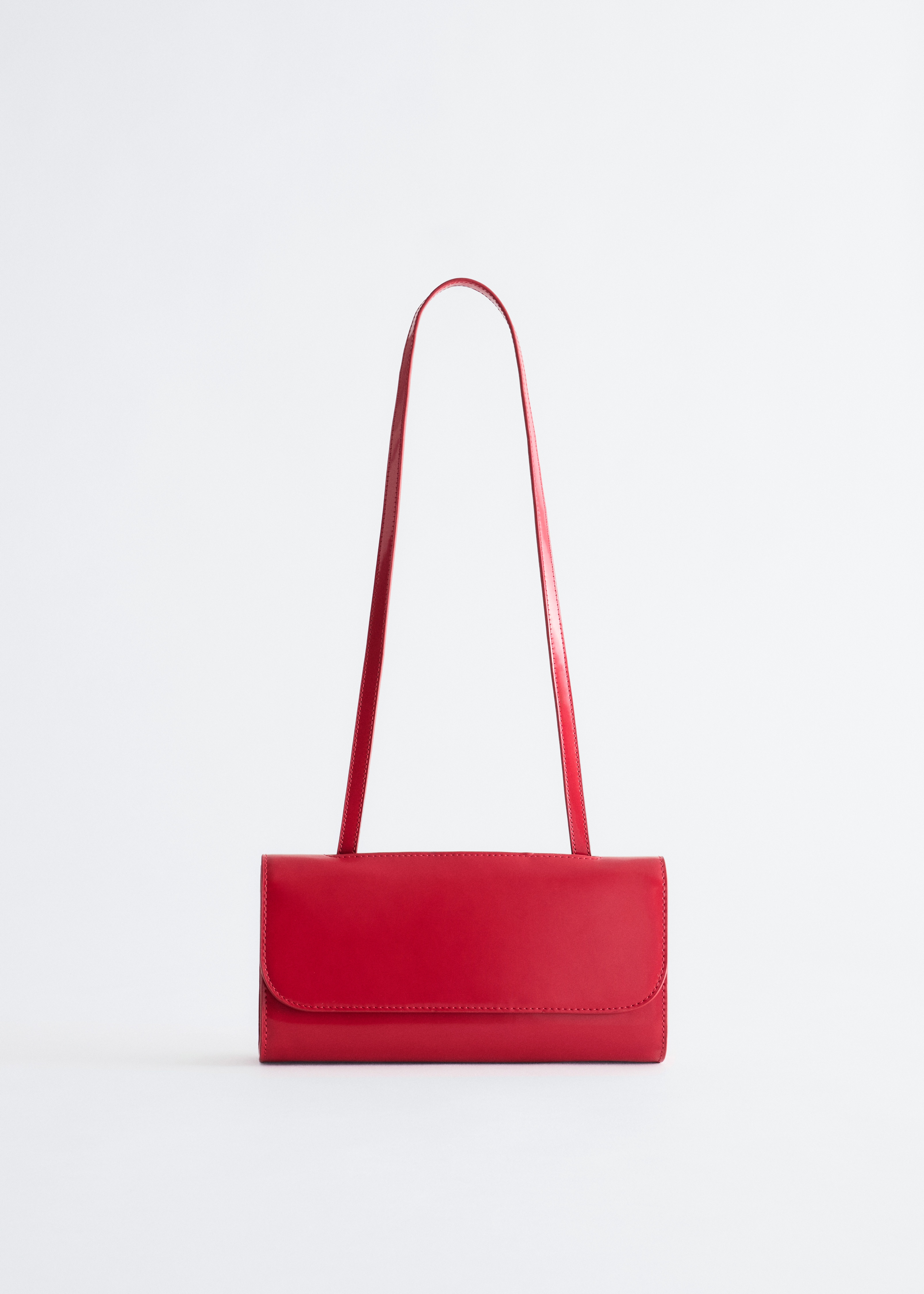 A structured, glossy red shoulder bag is being worn on the shoulder by the model. The bag features a clean, rectangular silhouette with a front flap and a slim shoulder strap, styled against a black sleeveless dress.