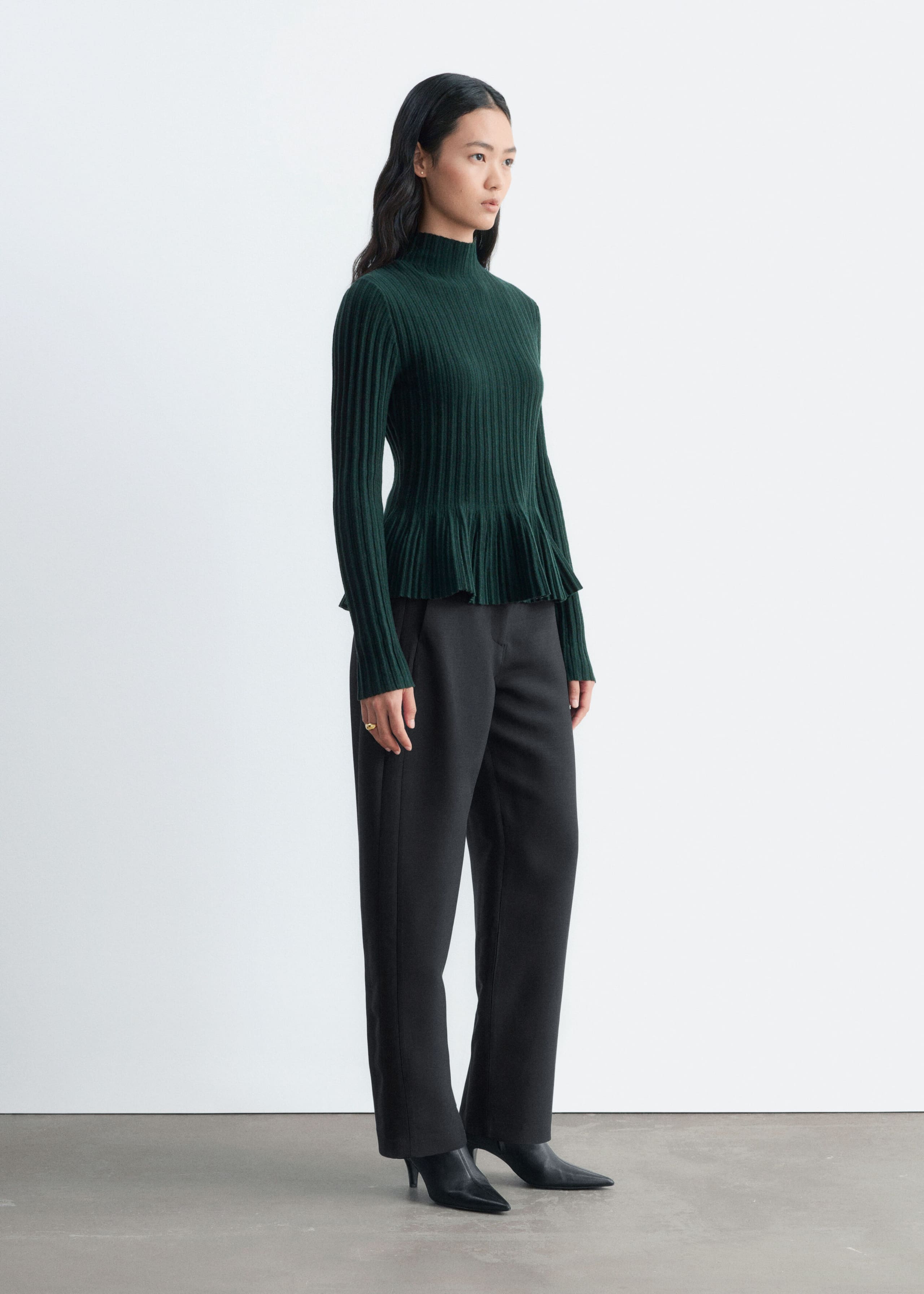 A dark green ribbed knitted top featuring a mock neck, long sleeves, and a flared peplum hem is being worn by the model. The top is styled with black trousers and black pointed-toe boots.