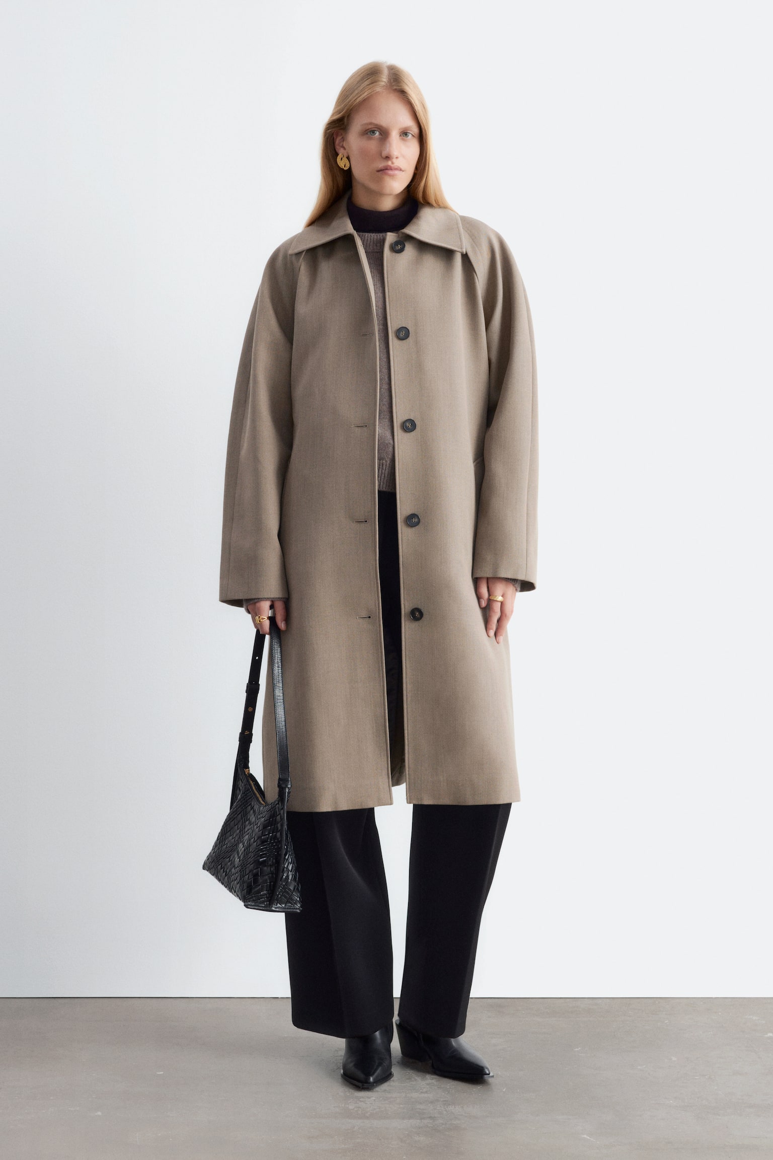 La modella indossa un trench in lana beige, portato aperto sopra un maglione in maglia grigio e pantaloni neri a gamba larga. Il cappotto, lungo al ginocchio, presenta un colletto classico e chiusure con bottoni scuri, mentre al fianco viene tenuta una borsa nera intrecciata in pelle. - 1