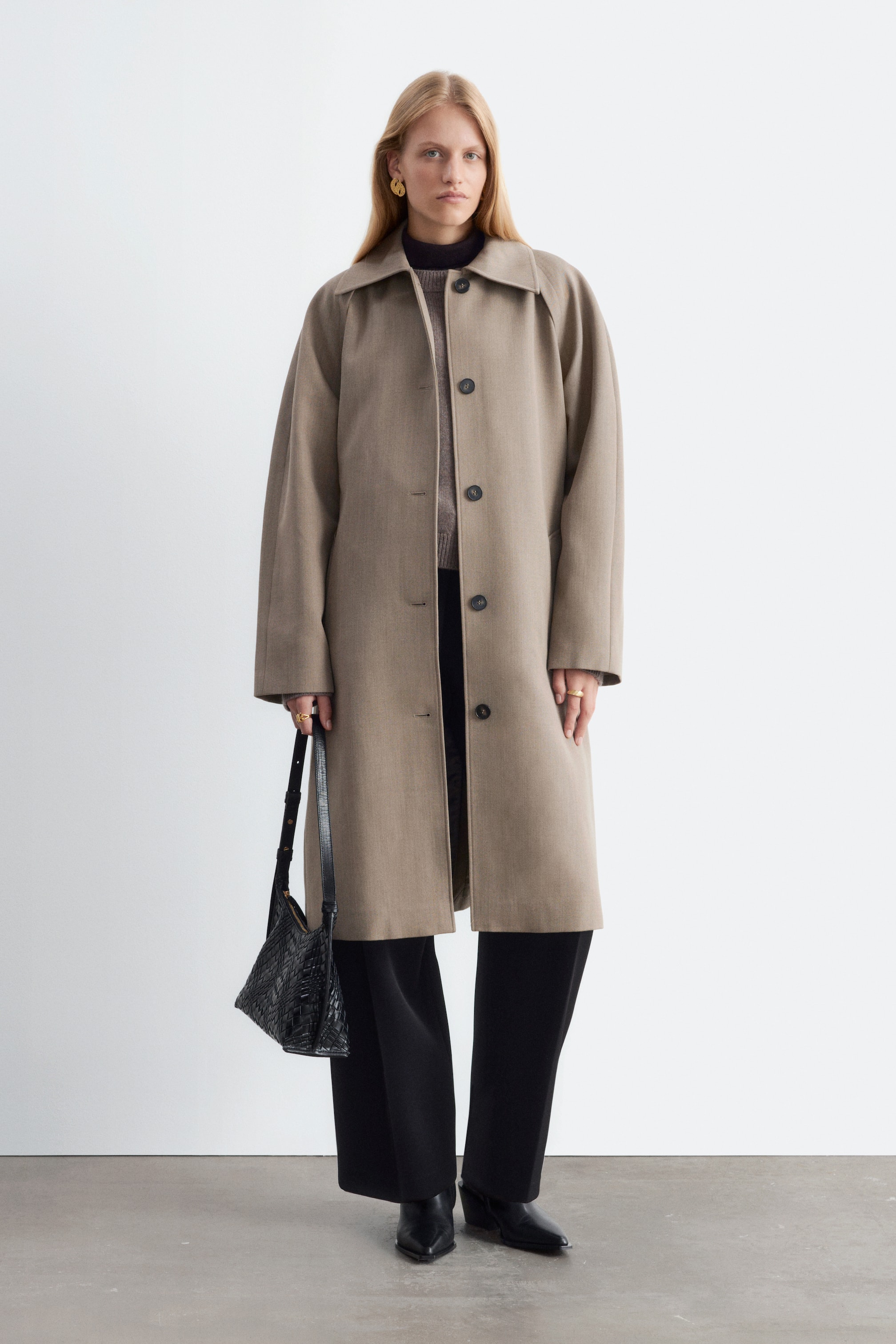 Größeres Bild ansehen: Ein beiger Woll-Trenchcoat wird vom Model getragen und offen über einem grauen Strickpullover und einer schwarzen Wide-Leg-Hose kombiniert. Der knielange Mantel verfügt über einen klassischen Kragen und dunkle Knopfverschlüsse, während seitlich eine schwarze, gewebte Lederhandtasche getragen wird.