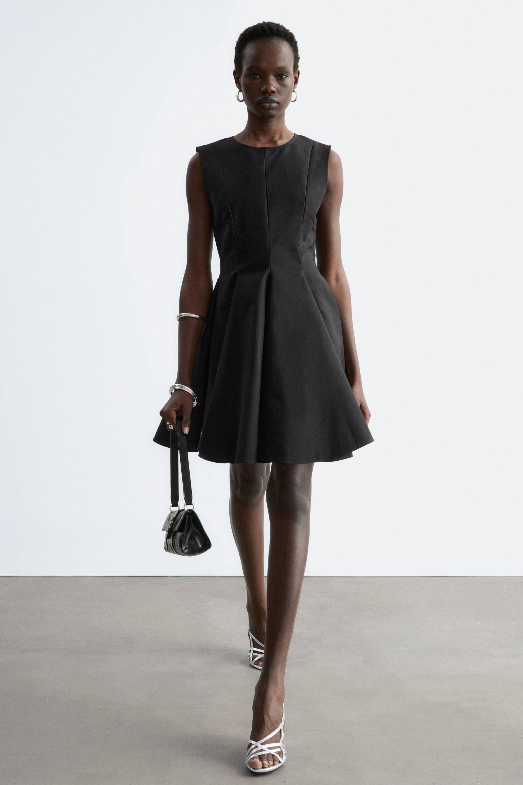 Vis større bilde: En svart ermeløs kjole med et figursydd liv med prinsessesømmer og en rund halslinje. Det fulle, A-formede skjørtet har dype folder som stråler ut fra midjen og ender over kneet.