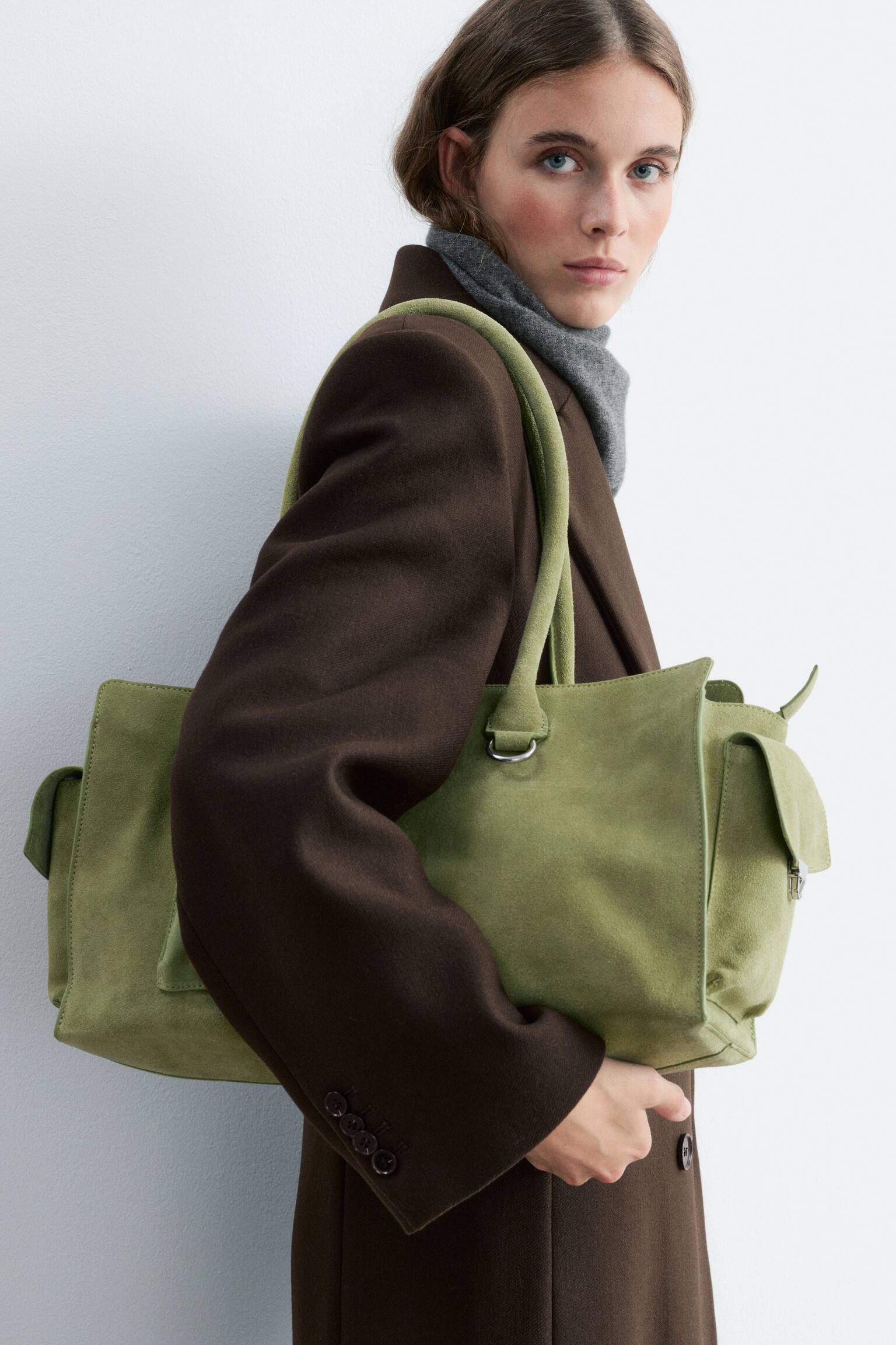 Un sac cabas en daim vert olive est prÃ©sentÃ© de face, avec une silhouette angulaire structurÃ©e et deux fines bandouliÃ¨res. Le design frontal comprend une poche verticale faÃ§on enveloppe avec des boutons-pression mÃ©talliques argentÃ©s Ã  gauche, un petit anneau en D mÃ©tallique argentÃ© Ã  droite, ainsi que des panneaux latÃ©raux Ã  rabat distinctifs. - 1