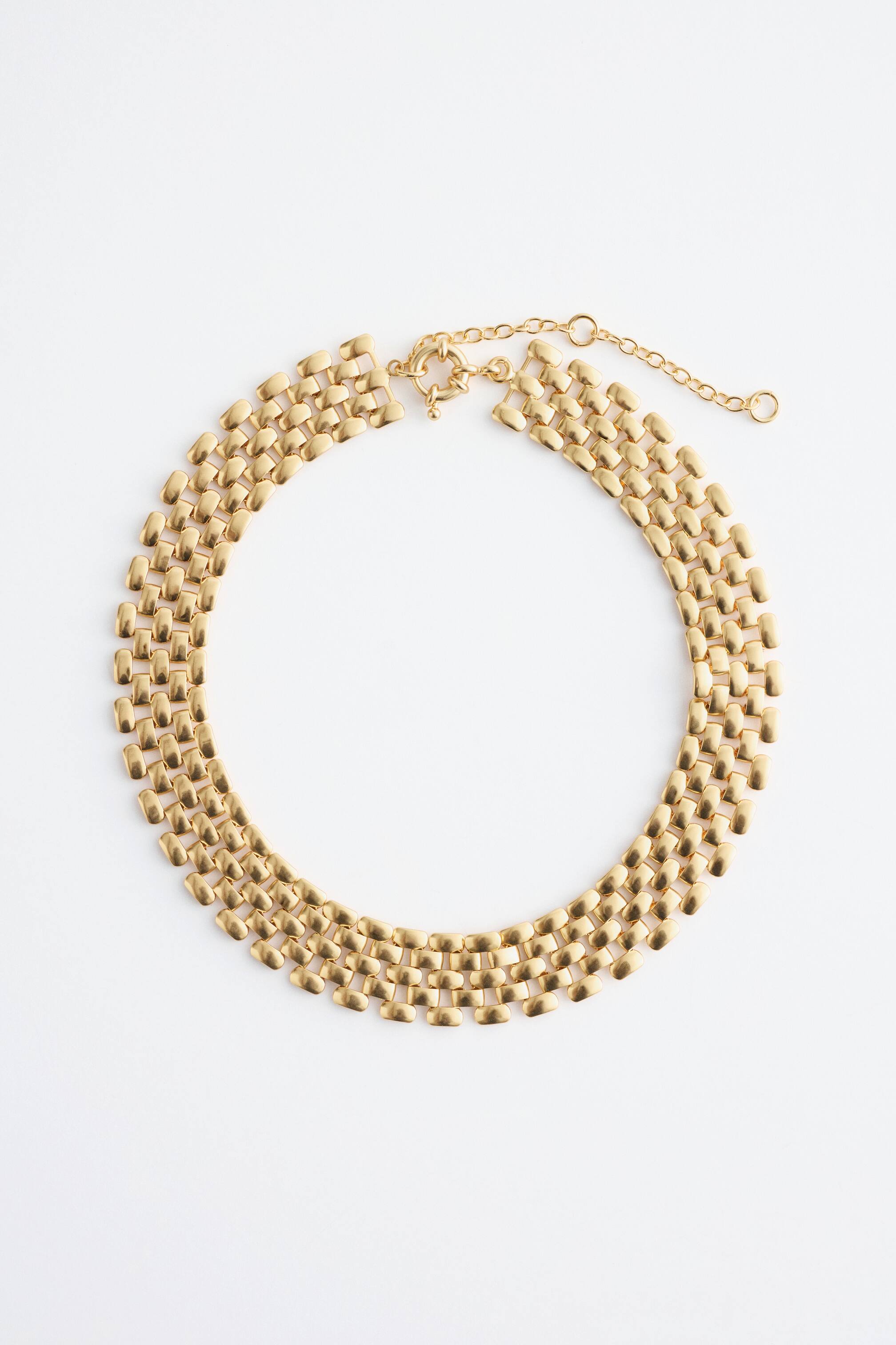 View larger image: A polished brass choker necklace is lying flat, featuring stacked square links forming a wide, architectural chain pattern. A toggle clasp and an extender chain are visible at one end, allowing for an adjustable fit.