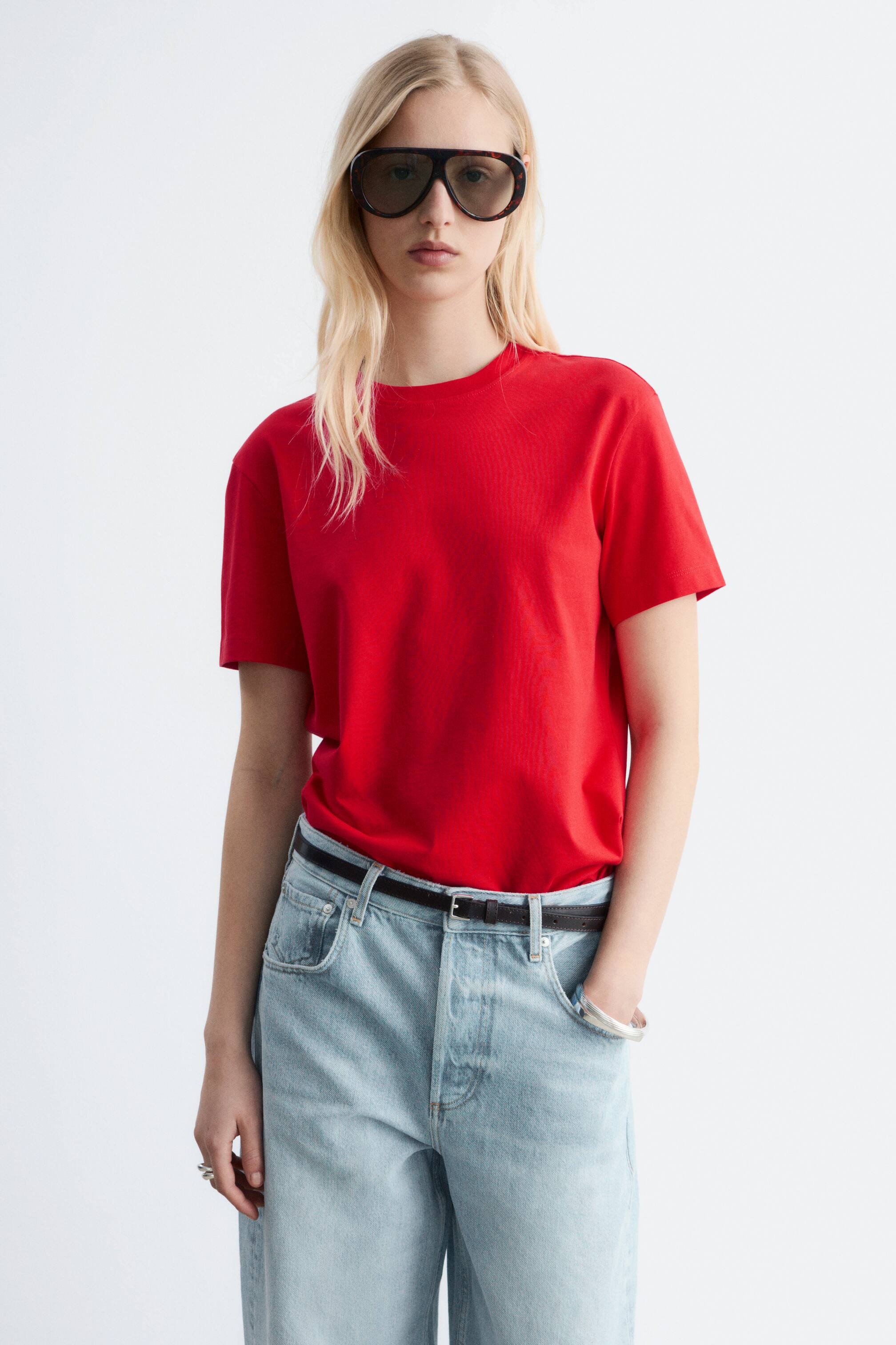 Größeres Bild ansehen: Ein rotes T-Shirt präsentiert sich mit einem Rundhalsausschnitt und kurzen Ärmeln, nach vorne gerichtet. Es wird in eine hellblaue Jeans gesteckt und mit einem schwarzen Gürtel fixiert.