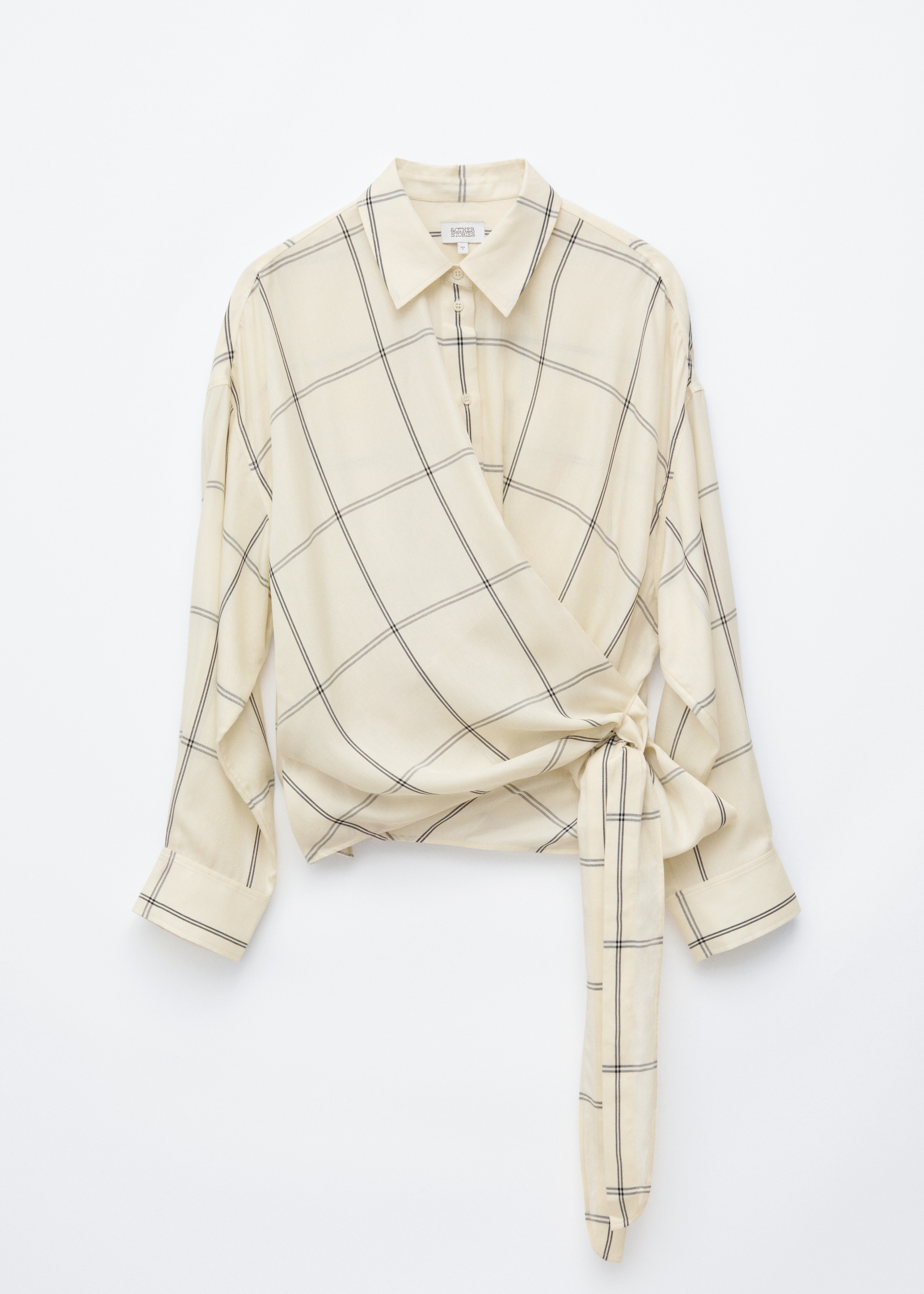 Une chemise en coton couleur crÃ¨me, Ã  motif quadrillÃ©, est portÃ©e par le mannequin. Elle prÃ©sente une coupe cache-cÅur nouÃ©e Ã  la taille, un col classique et des manches longues. La chemise est associÃ©e Ã  un pantalon noir taille haute.