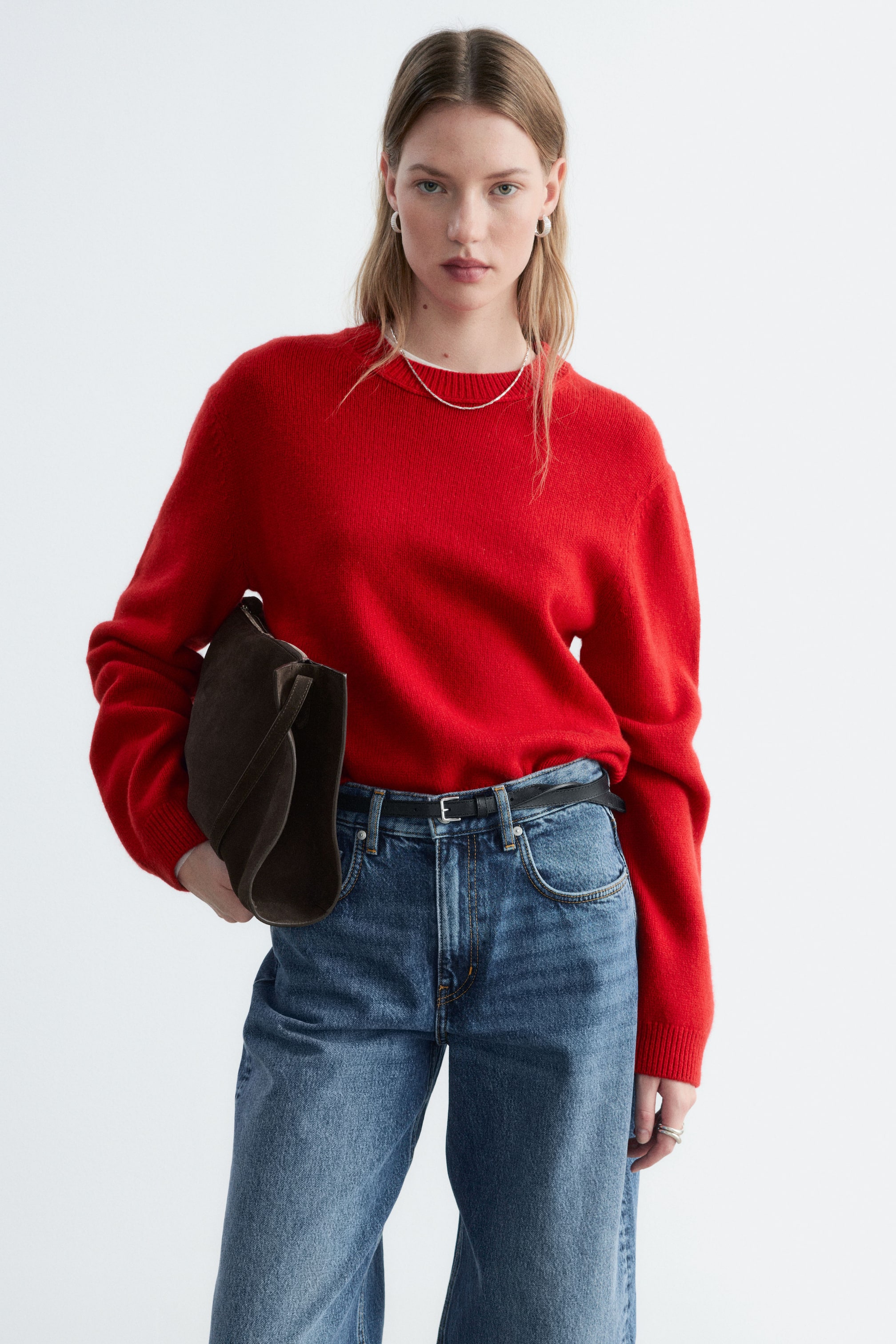 Visualizza l'immagine ingrandita: Un maglione in misto cashmere rosso con girocollo e polsini a coste è indossato dalla modella, infilato in jeans blu con una cintura nera. La modella tiene sotto un braccio una borsa in suede marrone scuro, appoggiandola al lato del maglione.