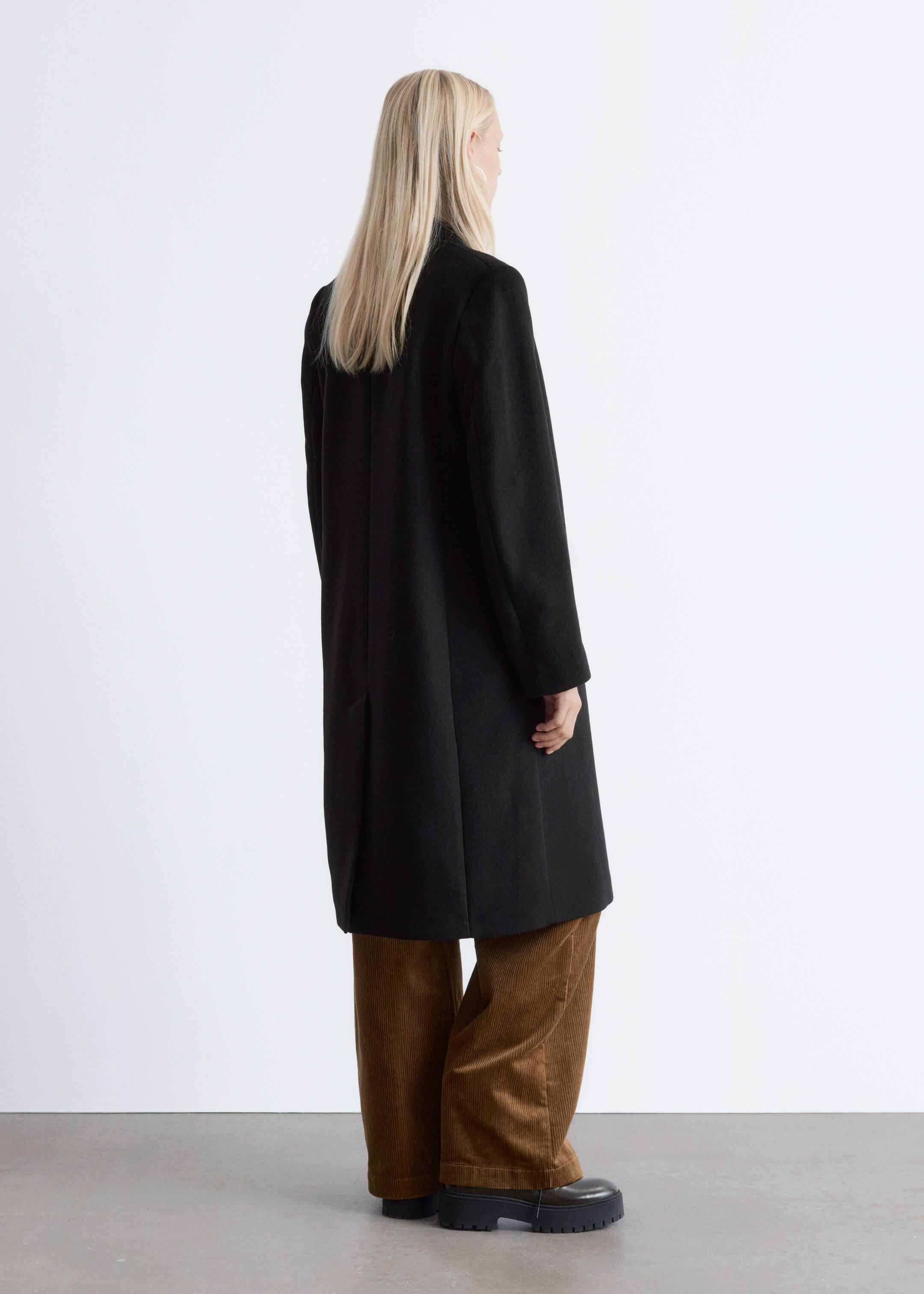 Agrandir l'image: Un manteau noir est présenté de dos, légèrement incliné vers la droite. Il présente une silhouette droite et ample avec des manches longues, un col montant et une couture centrale descendant le long du dos. Une fente subtile est visible près de l'ourlet, avec le manteau atteignant juste en dessous du genou.