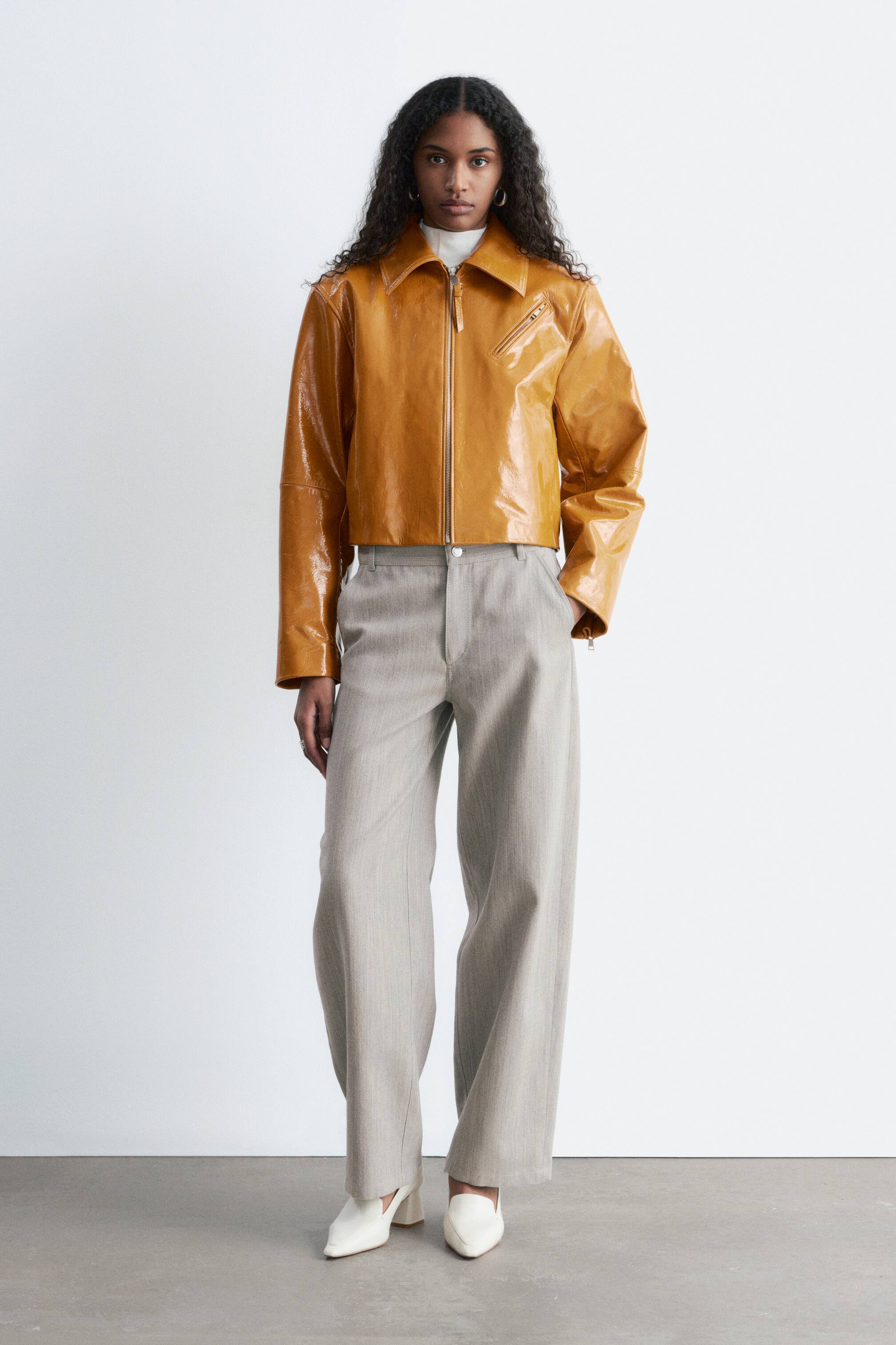 Größeres Bild anzeigen: Die kurz geschnittene Karamell-Patentjacke zeichnet sich durch einen weit gespreizten Kragen, einen durchgehenden Reißverschluss vorne und eine schräge Reißverschlusstasche auf der rechten Brust aus, präsentiert aus einer frontalen Perspektive. Sie wird über einem weißen Rollkragenpullover gestylt und mit hellgrauen, weit geschnittenen Hosen und cremeweißen, spitz zulaufenden Blockabsatzschuhen kombiniert.