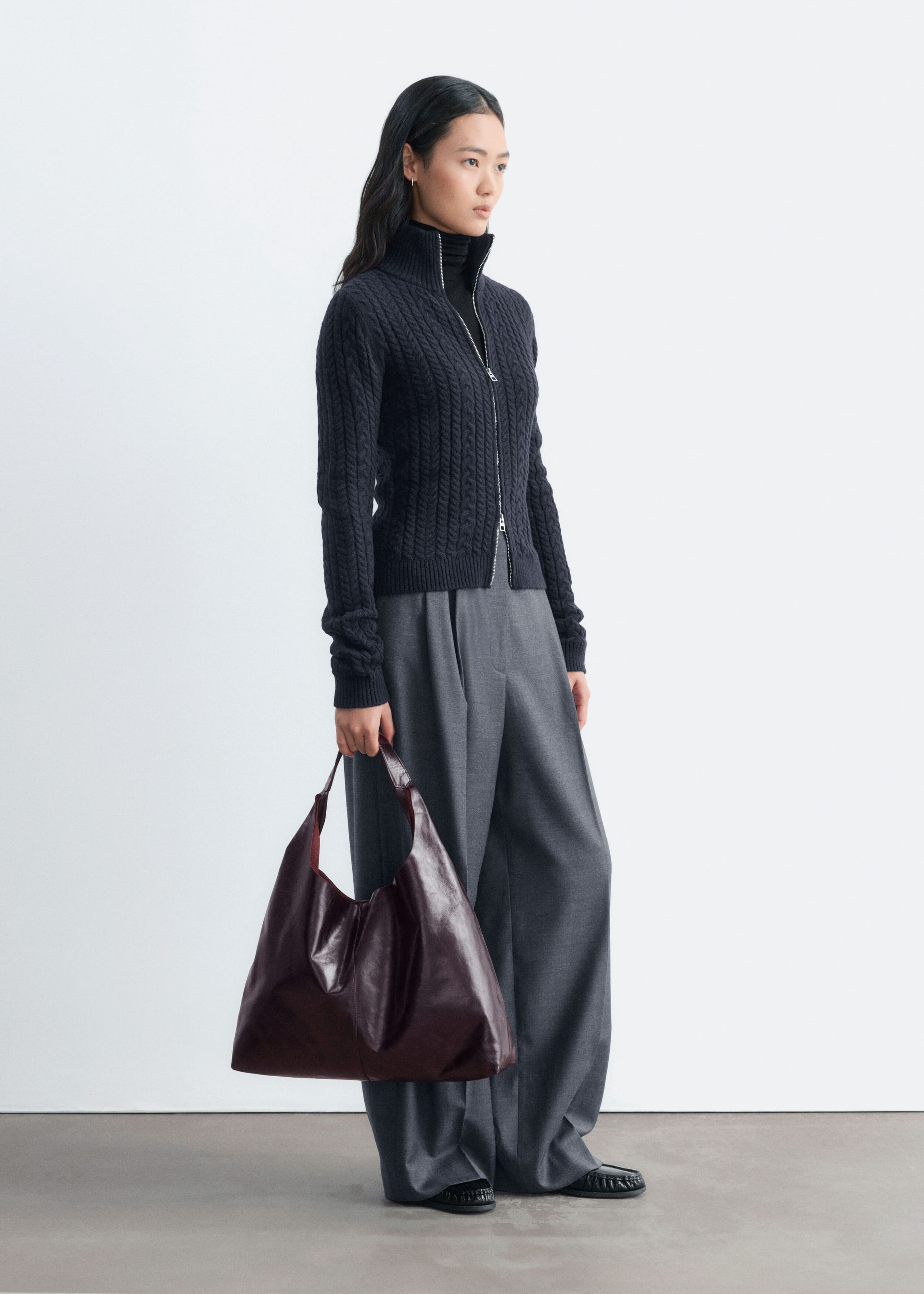 The model is wearing a dark grey cable-knit cardigan, layered over a black roll-neck top and paired with grey wide-leg trousers. The slim-fit cardigan features a high, double-layer ribbed collar and a silver front zip.
