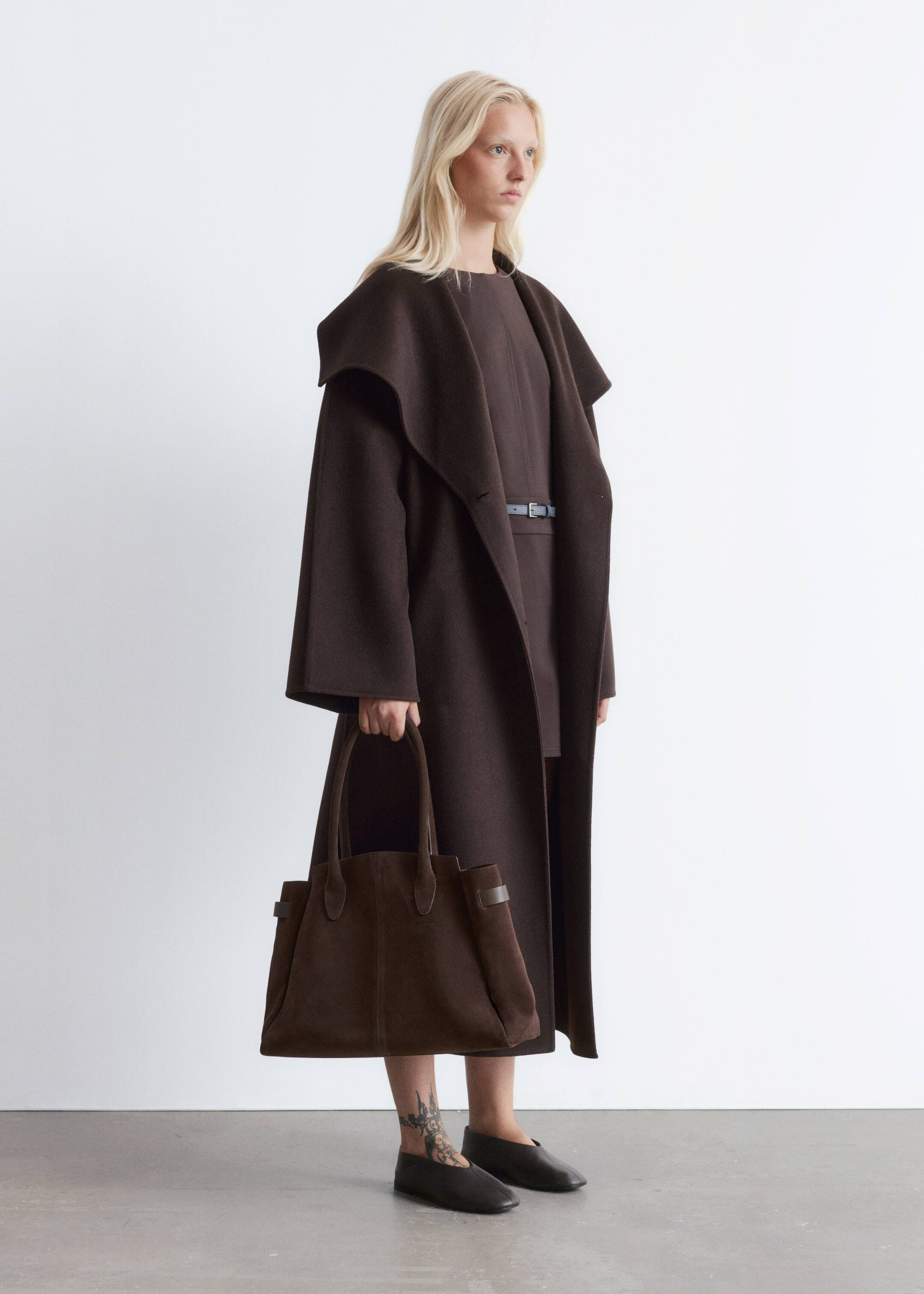 Visualizza immagine più grande: Un cappotto lungo marrone scuro con ampi revers drappeggiati è indossato, la sua vestibilità rilassata è visibile da un leggero profilo laterale. È stratificato su un vestito marrone scuro, stretto da una sottile cintura blu-grigio, mentre una borsa tote in camoscio marrone scuro con due manici superiori è tenuta nella mano destra.