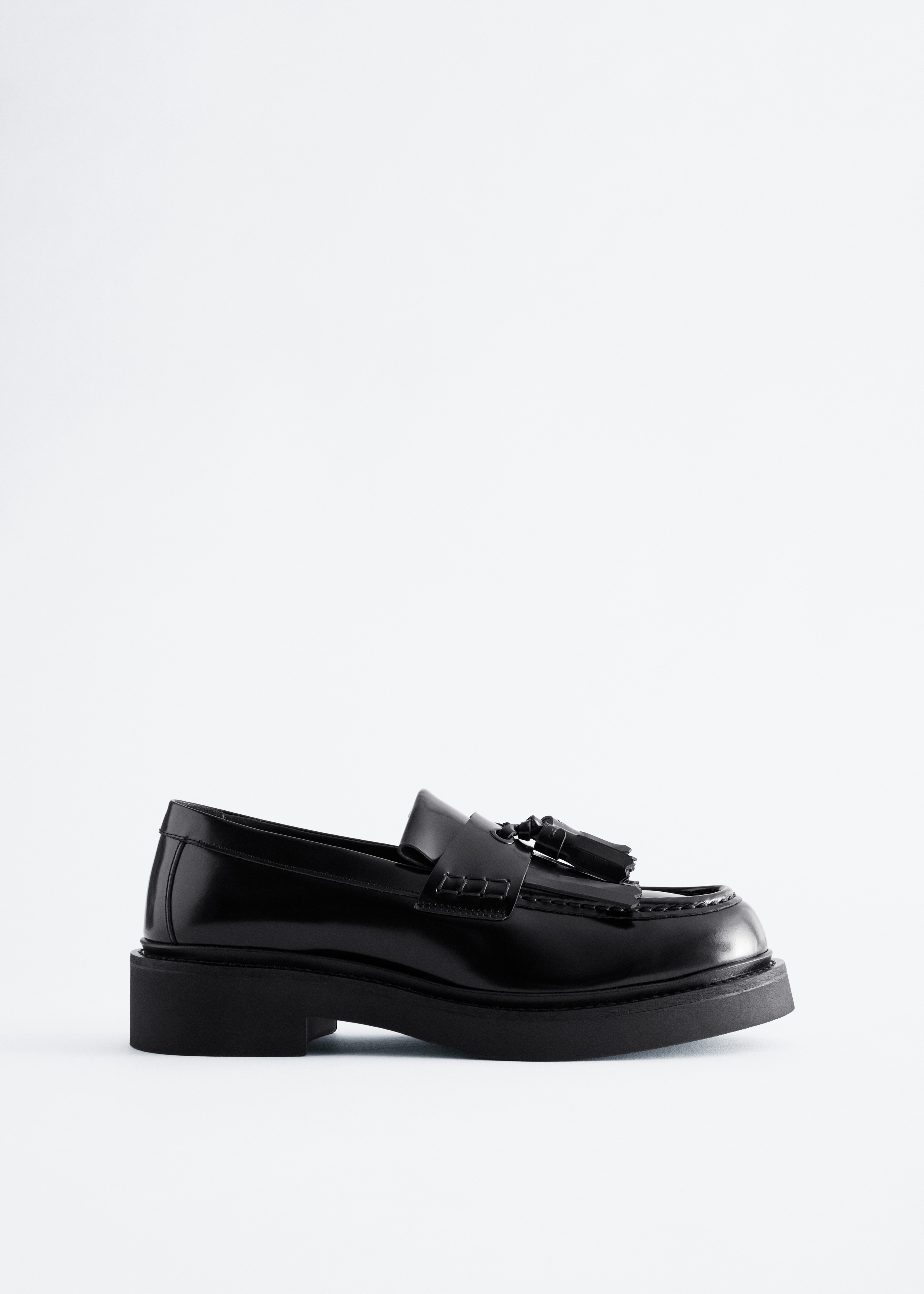 Il modello indossa mocassini in pelle nera lucida con frangia kiltie e nappine. Le scarpe con suola spessa sono abbinate a pantaloni a quadri scuri dalla texture marcata.