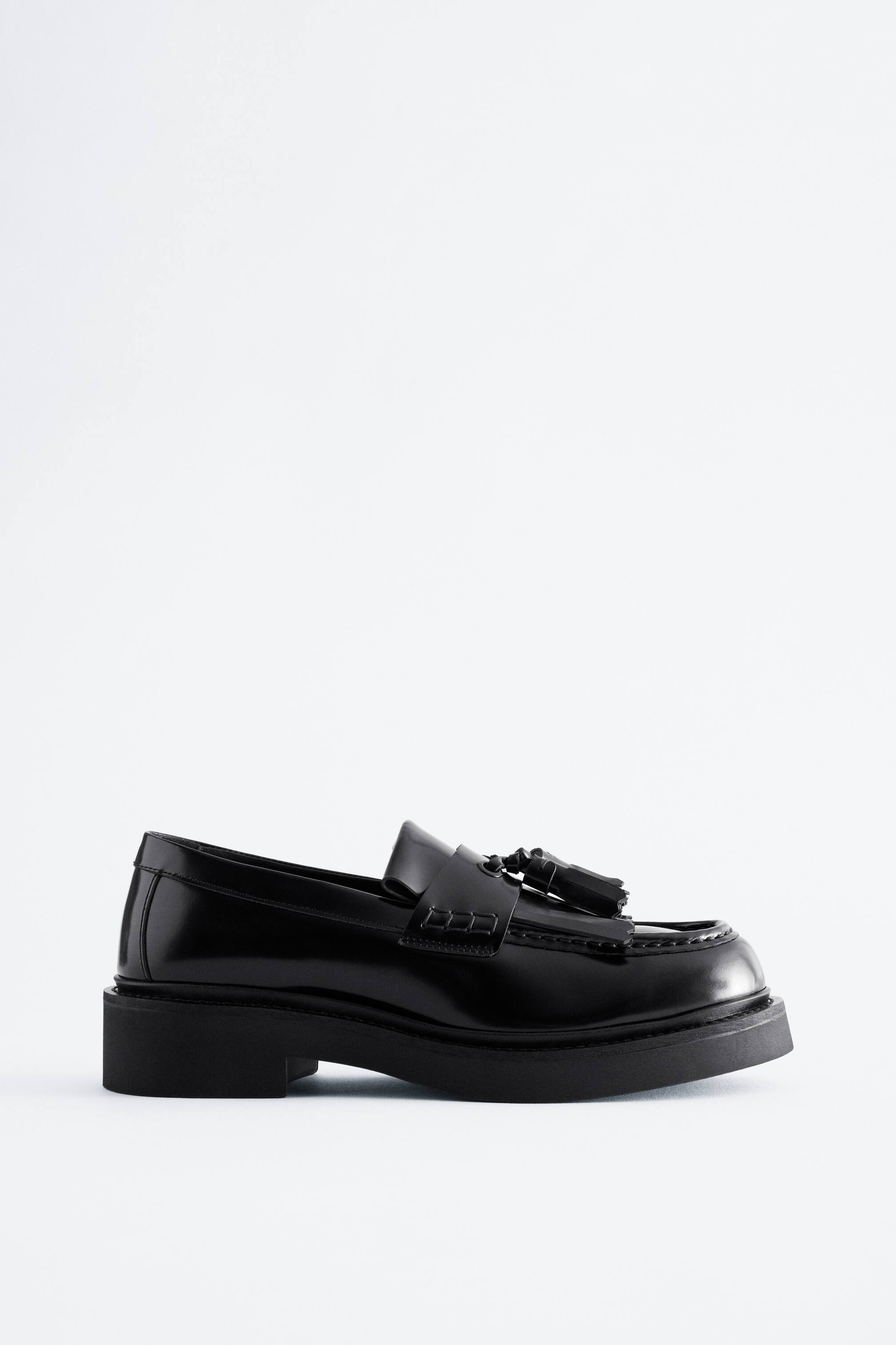 Ver imagen más grande: Un mocasín de cuero negro pulido aparece en vista de perfil, mostrando una gruesa suela negra y un tacón bajo de bloque. El zapato presenta borlas decorativas y un detalle de flecos kiltie sobre el empeine, junto con costuras moc-toe visibles.