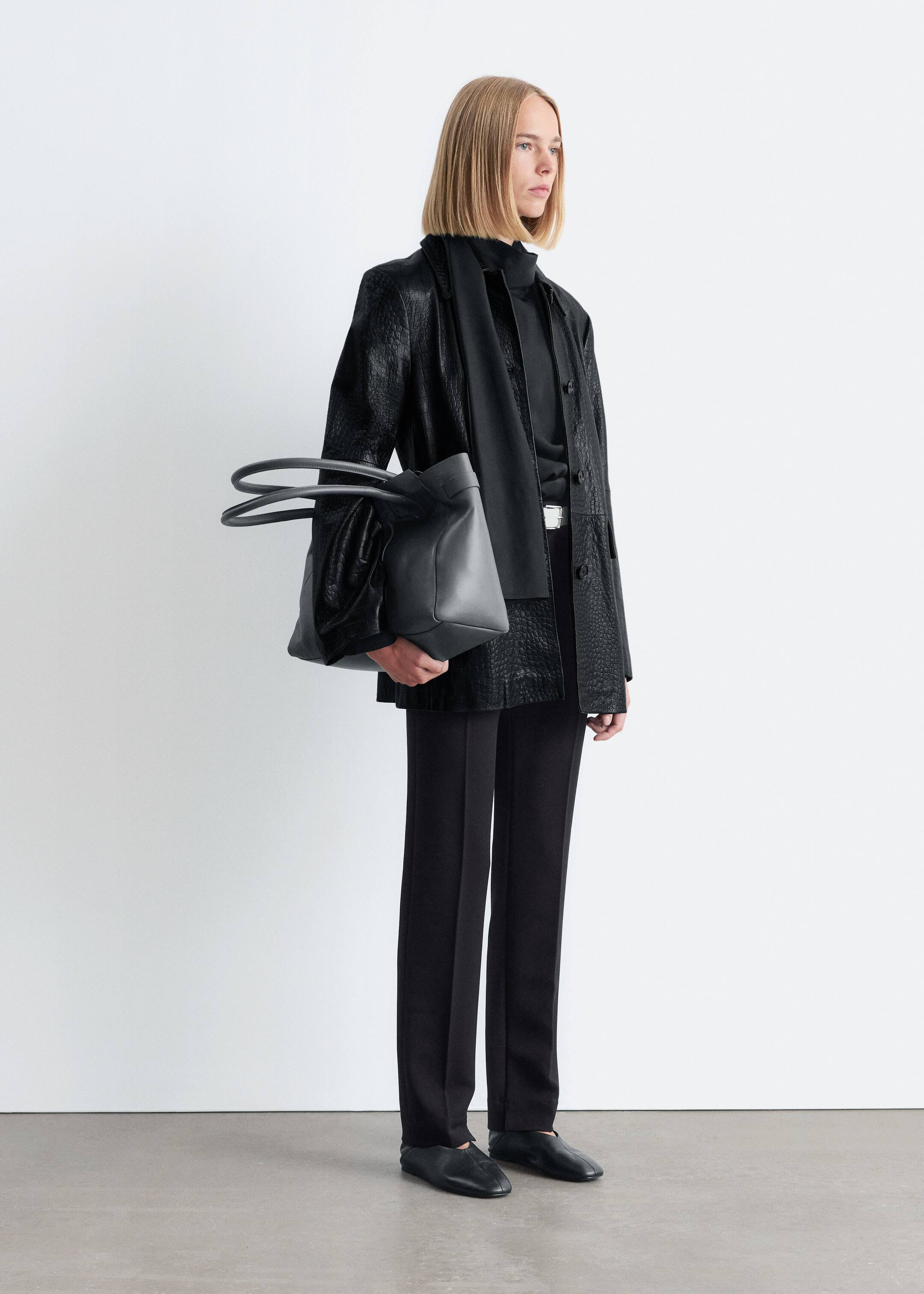Größeres Bild ansehen: Ein schwarzes Modeensemble wird aus einer 3/4-Frontansicht präsentiert. Es verfügt über eine offene Jacke mit einem geprägten Muster, die über einem schwarzen Oberteil mit hohem Kragen und gerade geschnittenen Hosen getragen wird. Ein schwarzer Schal ist um den Hals drapiert, schwarze Slipper vervollständigen den Look, und eine dunkelgraue weiche Ledertasche mit abgerundeten Griffen wird in der linken Hand des Models gehalten, leicht nach vorne geneigt.