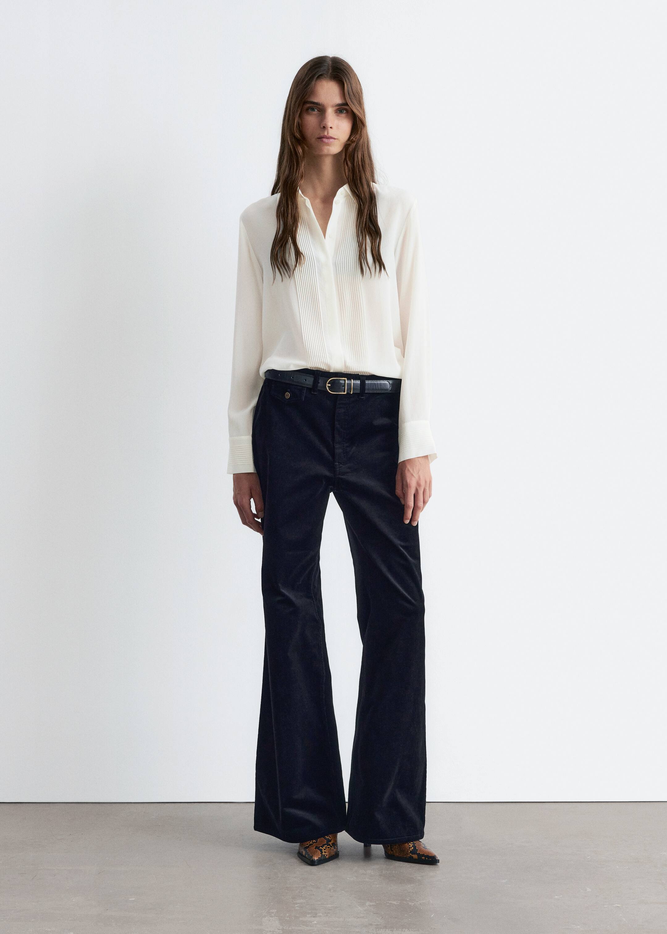 Grotere afbeelding bekijken: Een witte blouse met lange mouwen, met verticale pintucks langs de voorkant en horizontaal geplooide manchetten, wordt gedragen. Het wordt gecombineerd met zwarte fluwelen broek met wijde pijpen, die worden aangesnoerd door een zwarte riem met een gouden gesp en vallen over puntige slangenleren laarzen.