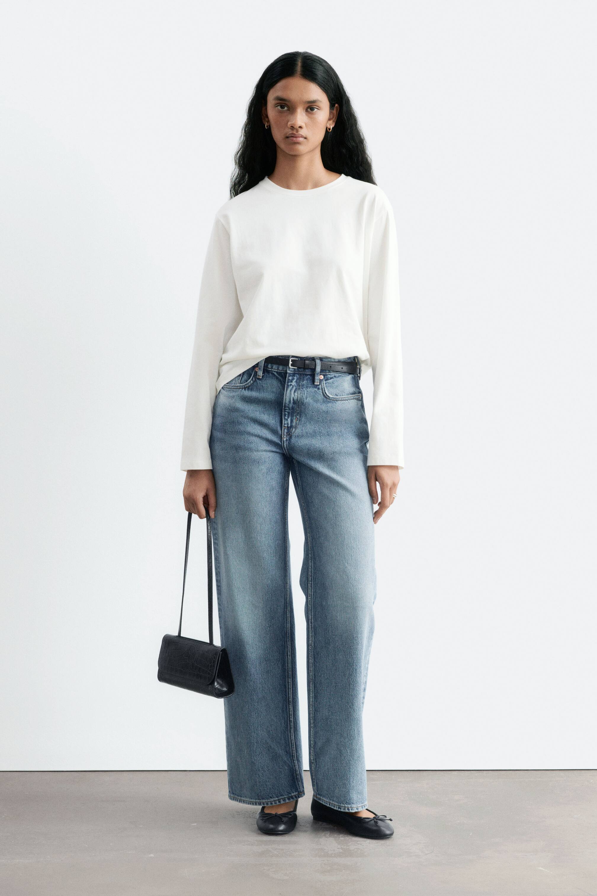 Se større billede: Modelen bærer blå jeans med lige ben, mellemhøj talje og lys vask. Jeansene er stylet med en nedstukket hvid langærmet top, et tyndt sort bælte og sorte ballerinasko, mens der bæres en lille sort håndtaske.