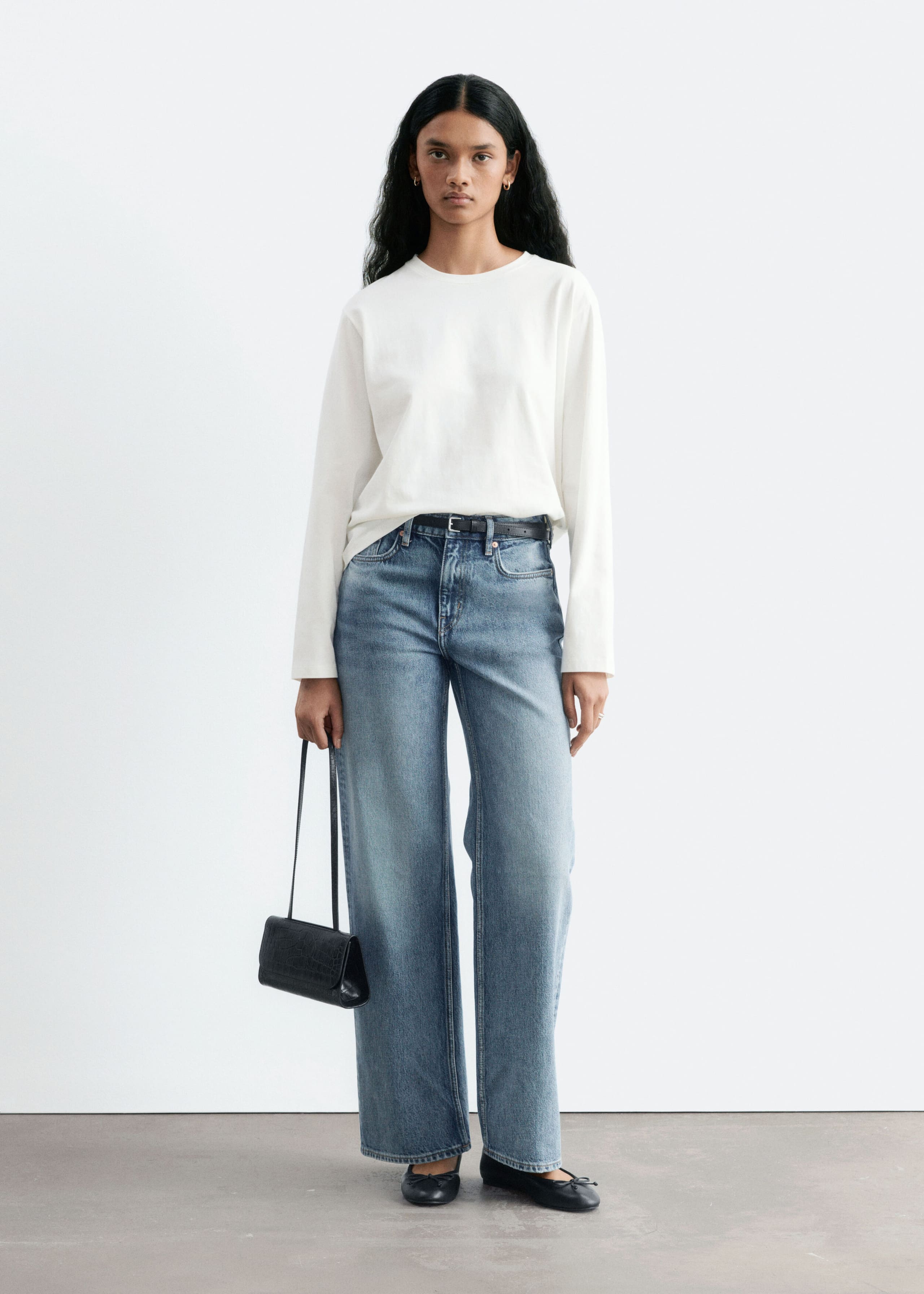 The model is wearing straight-leg blue jeans with a mid-rise waist and a light wash. The jeans are styled with a tucked-in white long-sleeved top, a slim black belt, and black ballet flats, while carrying a small black handbag.