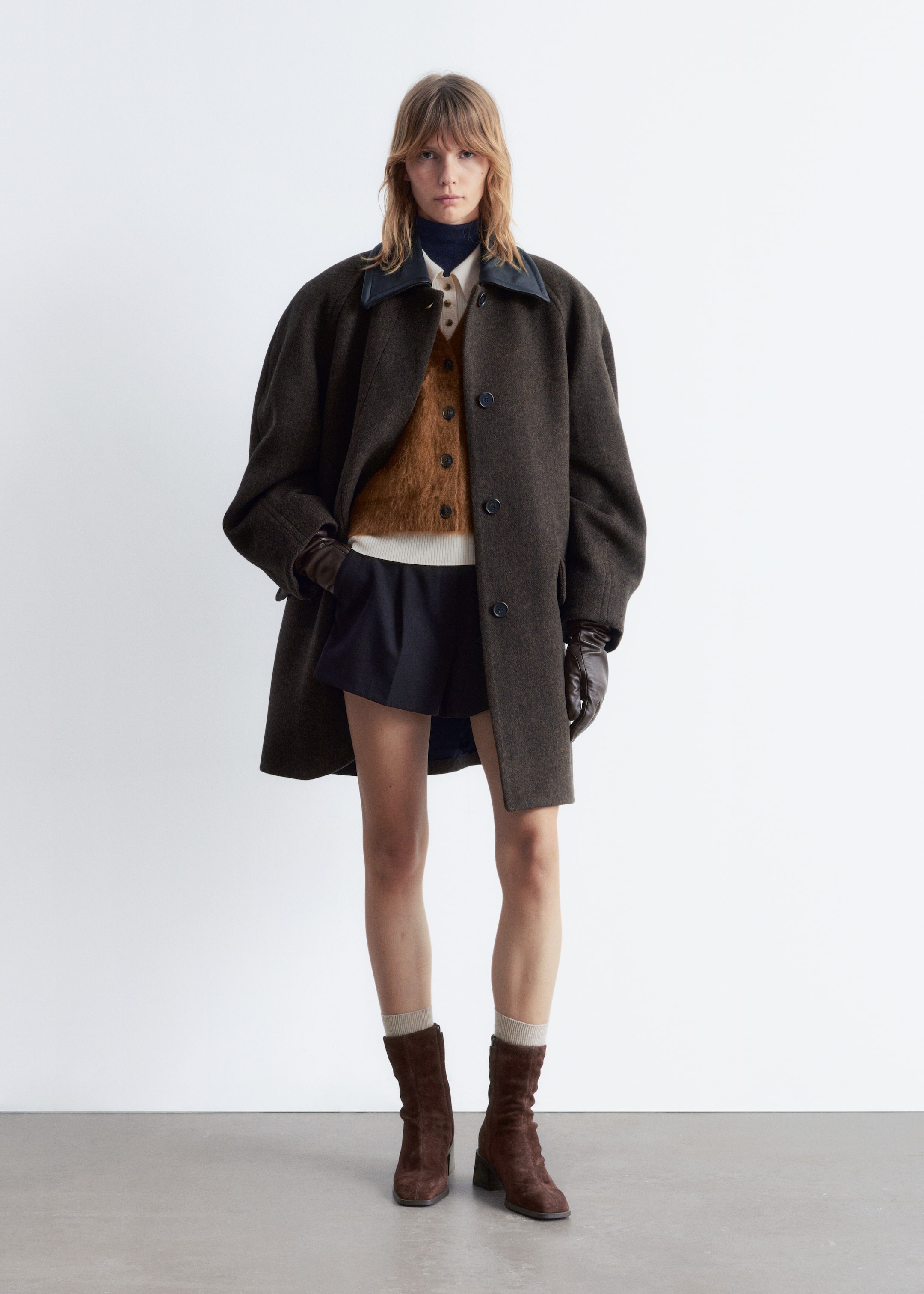 Agrandir l'image: Un manteau brun foncé est exposé de face, mettant en avant un col pointu foncé contrastant et une fermeture boutonnage simple avec des boutons foncés. Sa coupe ample met en valeur son drapé sur des vêtements superposés, avec des mains dans ses poches latérales, portant des gants en cuir brun foncé.