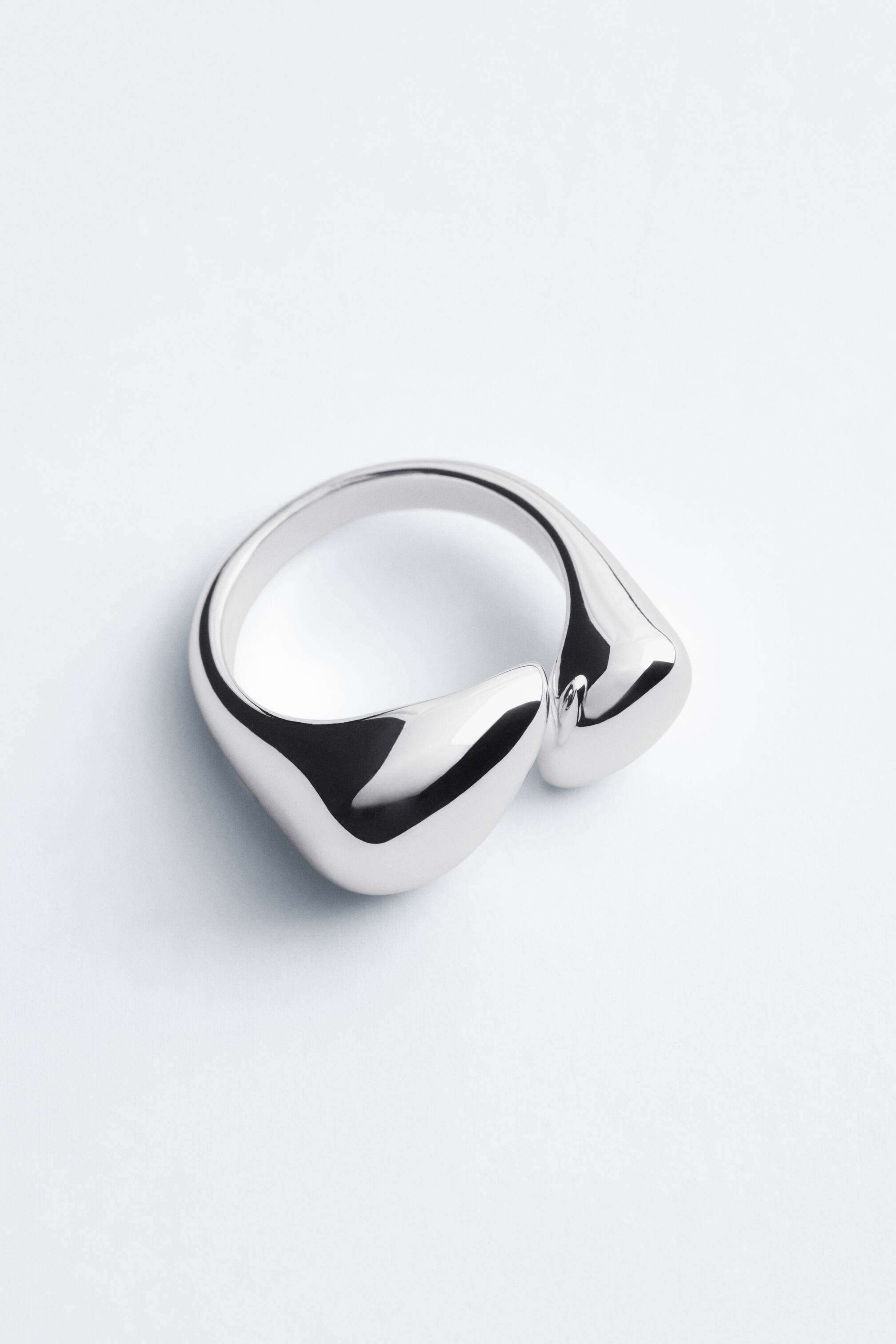 Grösseres Bild ansehen: Ein polierter, silberfarbener Ring liegt auf einer hellen Oberfläche und zeigt ein offenes, skulpturales Design mit fliessenden, bauchigen Enden, die sich vorne einander zuneigen.