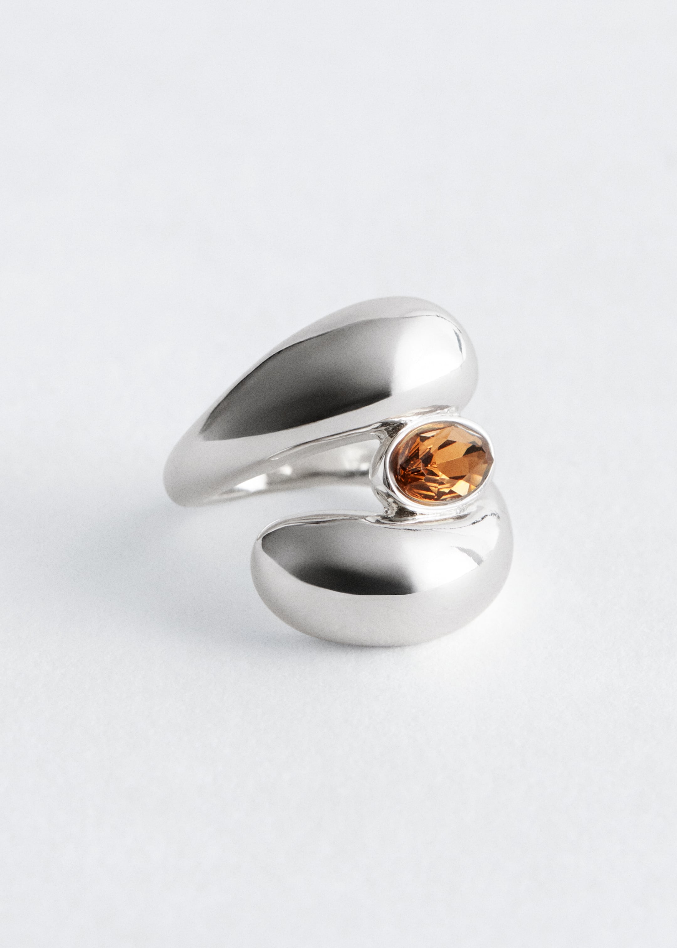 Agrandir l'image: Une bague polie de ton argenté est présentée de FACE, son design enveloppant massif avec des bords arrondis. Un cristal facetté de couleur ambre en forme ovale est maintenu en son centre, là où les deux extrémités du design enveloppant se rejoignent.