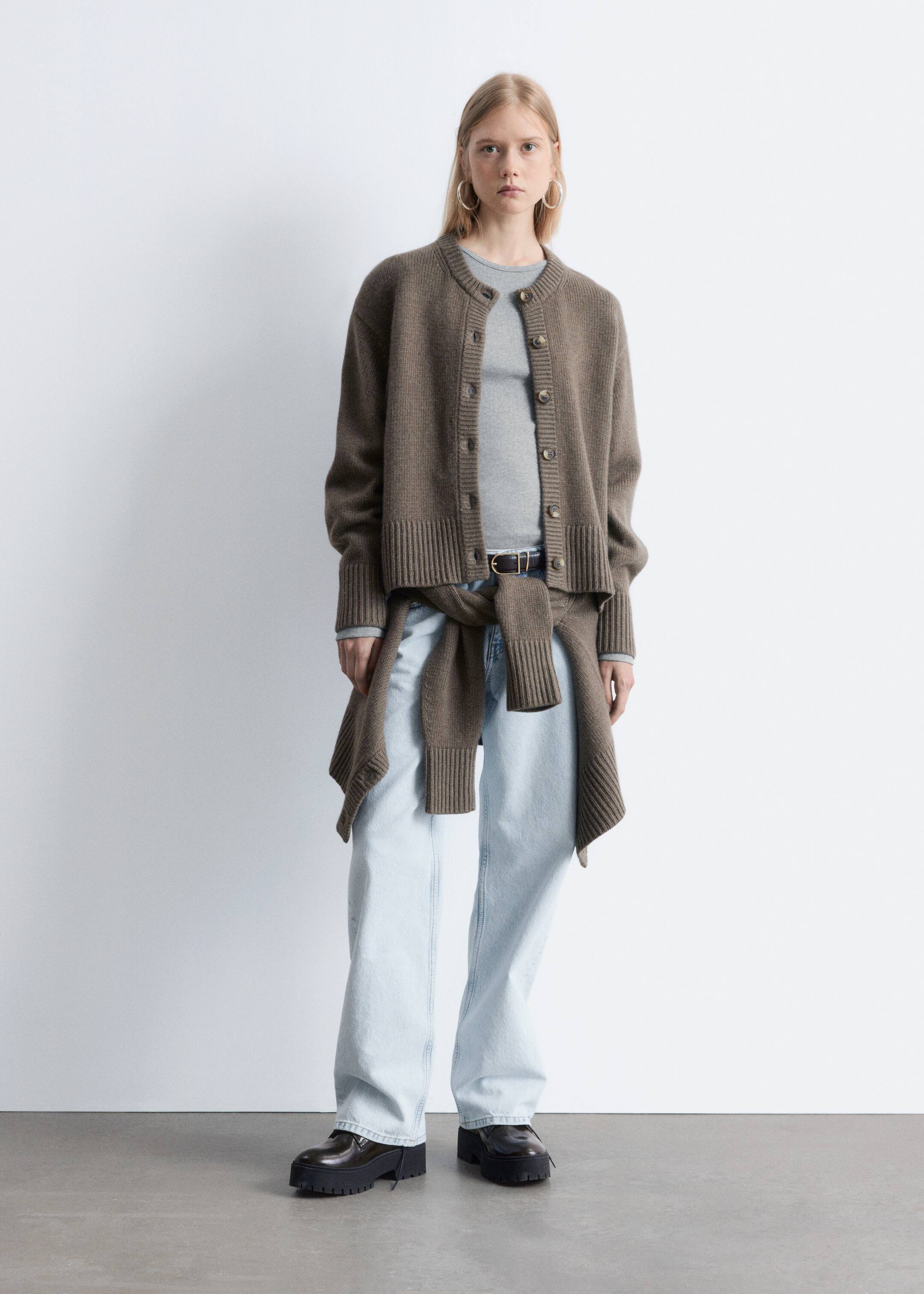 Agrandir l'image: Un cardigan en tricot brun est porté par-dessus un haut gris clair, face vers l'avant. Il est conçu avec un col rond, des manches longues, et une fermeture à boutons sur le devant avec plusieurs petits boutons bruns et ronds. Le vêtement à la coupe décontractée s'étend jusqu'aux hanches, mettant en valeur des ourlets et poignets côtelés, et un vêtement en tricot brun assorti est noué autour de la taille.