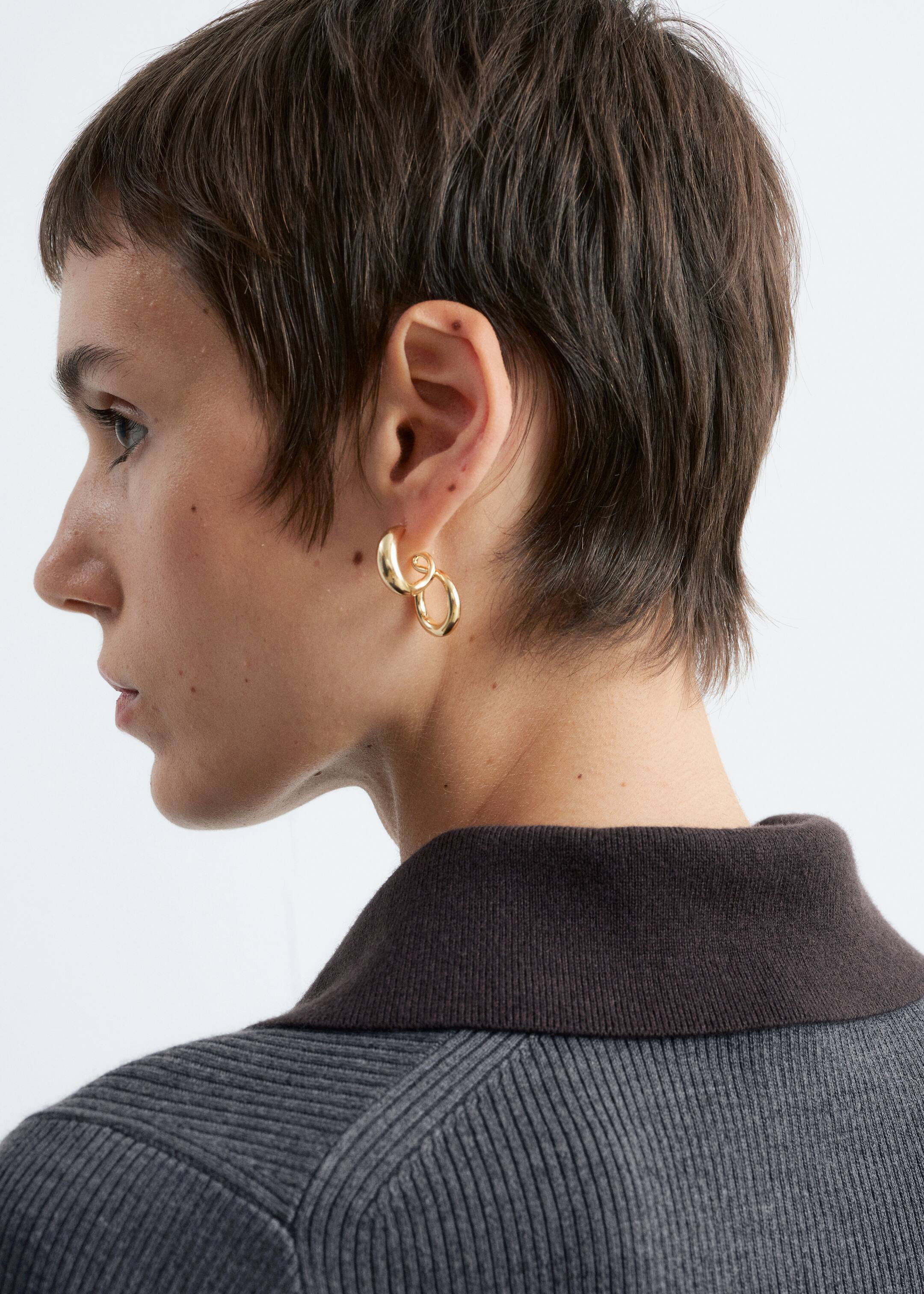 Agrandir l'image: Des boucles d'oreilles dorées pendent d'un lobe d'oreille, présentant deux éléments sculpturaux interconnectés. Un anneau solide et arrondi s'emboîte avec une spirale ouverte et allongée, tous deux vus de profil juste au-dessus du col marron foncé d'un vêtement gris côtelé.