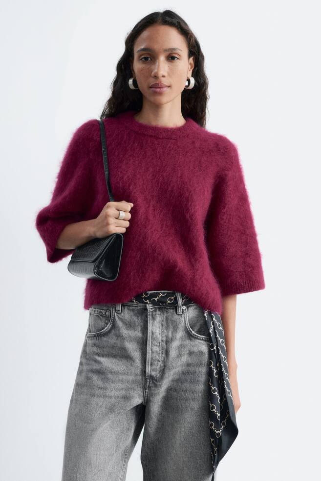 Een zachte, bessenkleurige gebreide T-shirt wordt gedragen door het model, met een geborstelde mohairstructuur, een ronde hals en losse mouwen tot op de elleboog. De T-shirt is gestyled met een high-waisted grijze jeans en een zwarte schoudertas die onder de arm wordt gedragen. - 1