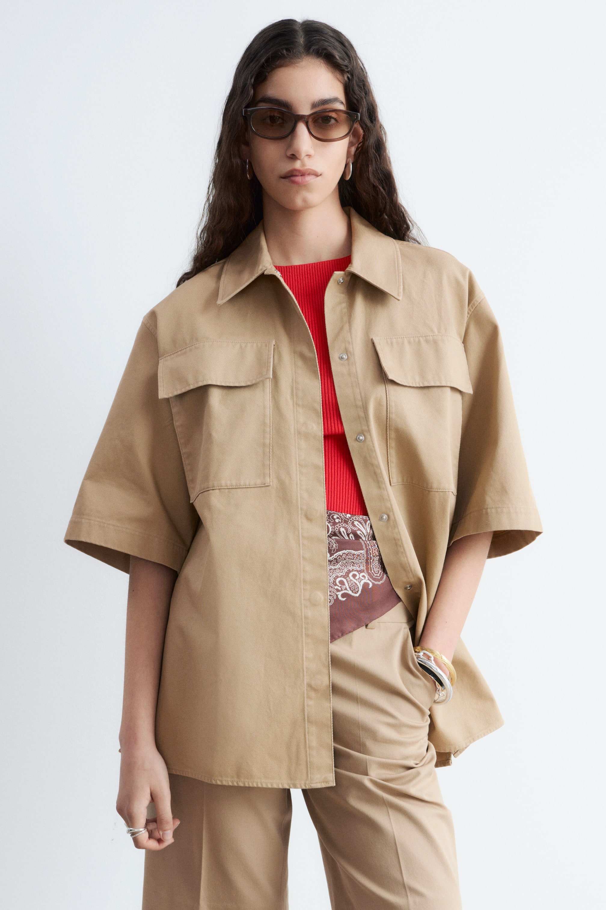 Nagyobb kép megtekintése: Egy bézs, túlméretezett rövid ujjú ing látható hegyes gallérral, két zsebfedeles mellzsebbel és elöl ezüst patentos gombolópánttal. Nyitva viselik egy piros felső és egy barna mintás kendő fölött, és kiegészíti a hozzá illő bézs nadrágot.