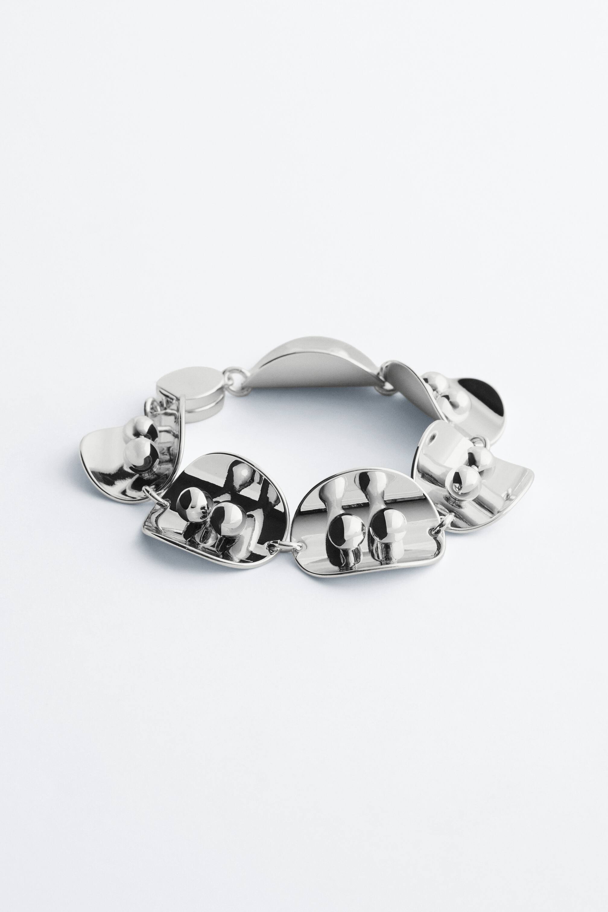 View larger image: A silver-toned bracelet lies flat, featuring multiple linked segments. Some segments are smooth and curved, while others are curved and display raised, rounded, elongated shapes on their surface.