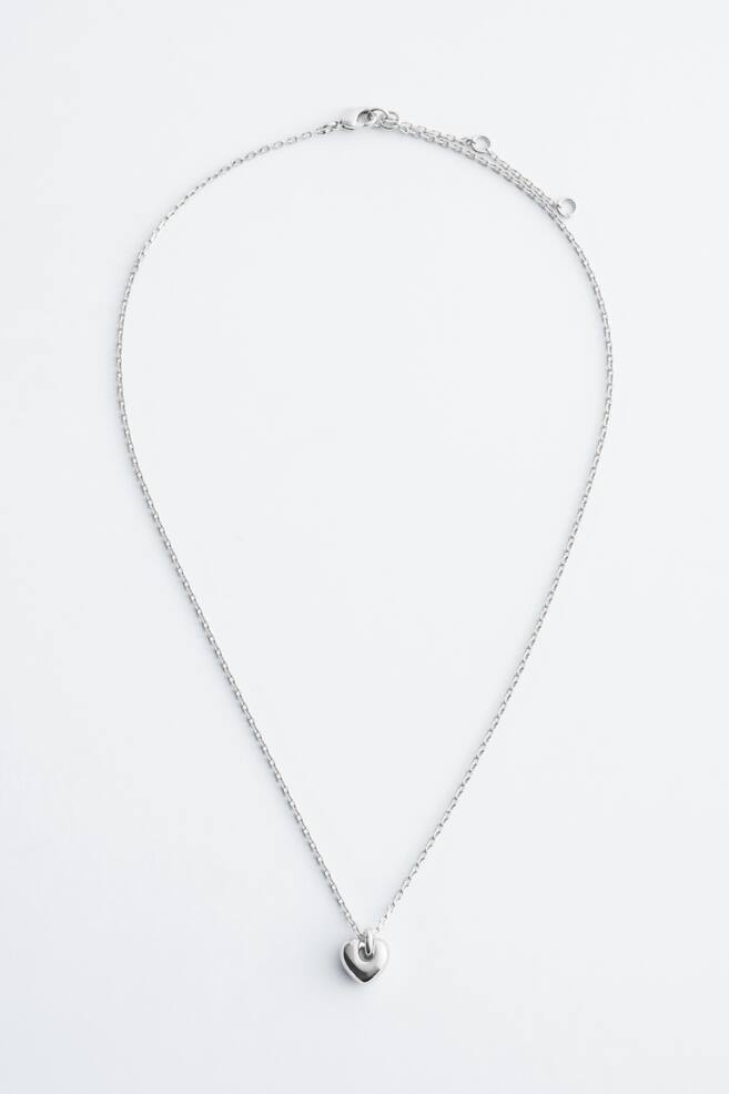 A silver-toned necklace featuring a delicate link chain, a polished, chunky heart pendant, and an adjustable extender chain with a trigger clasp visible at the back. - 1