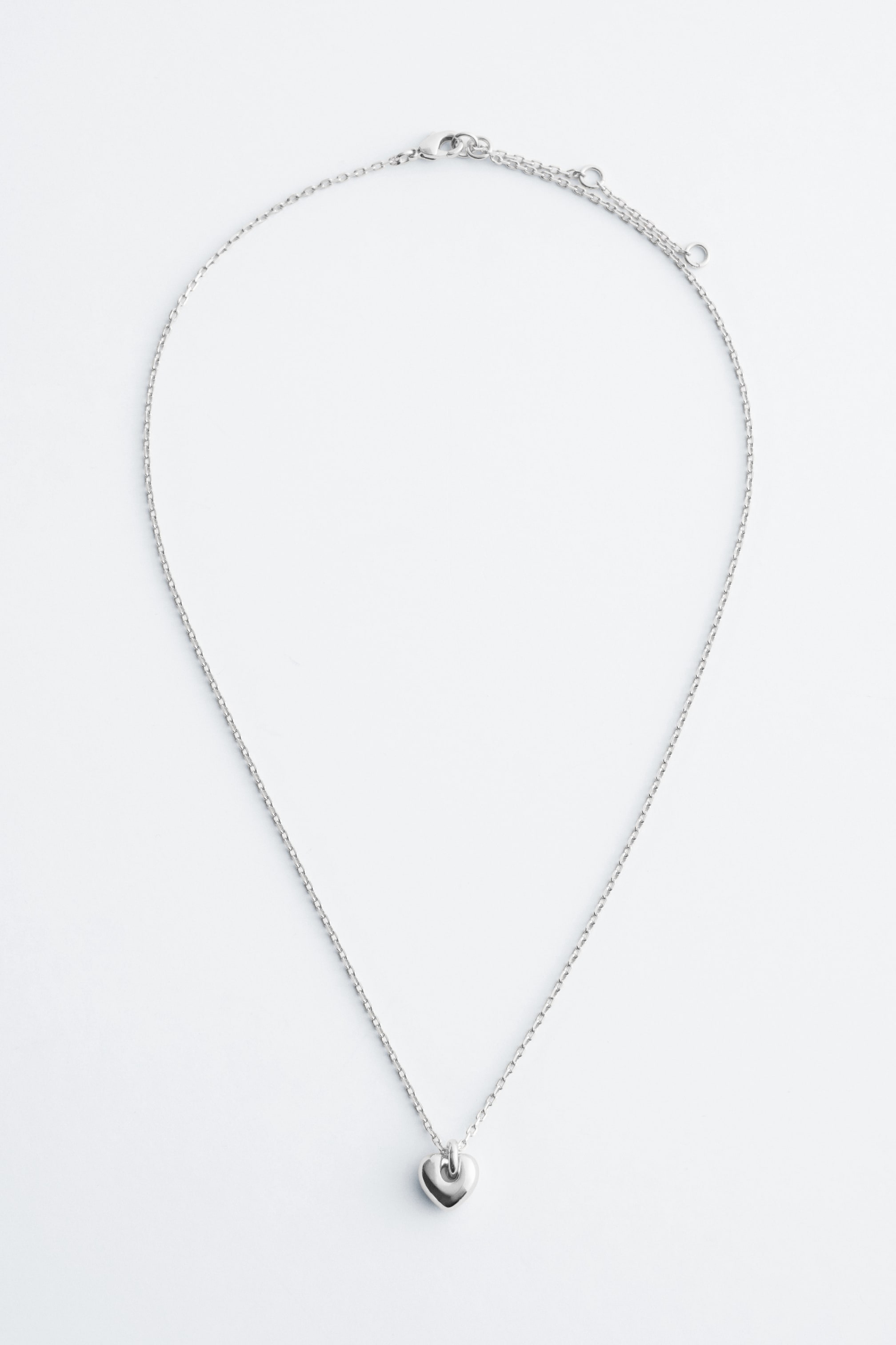 Visa större bild: Ett silverfärgat halsband med en tunn länkkedja, ett polerat, kraftigt hjärthänge och en justerbar förlängningskedja med en synlig karbinhake baktill.