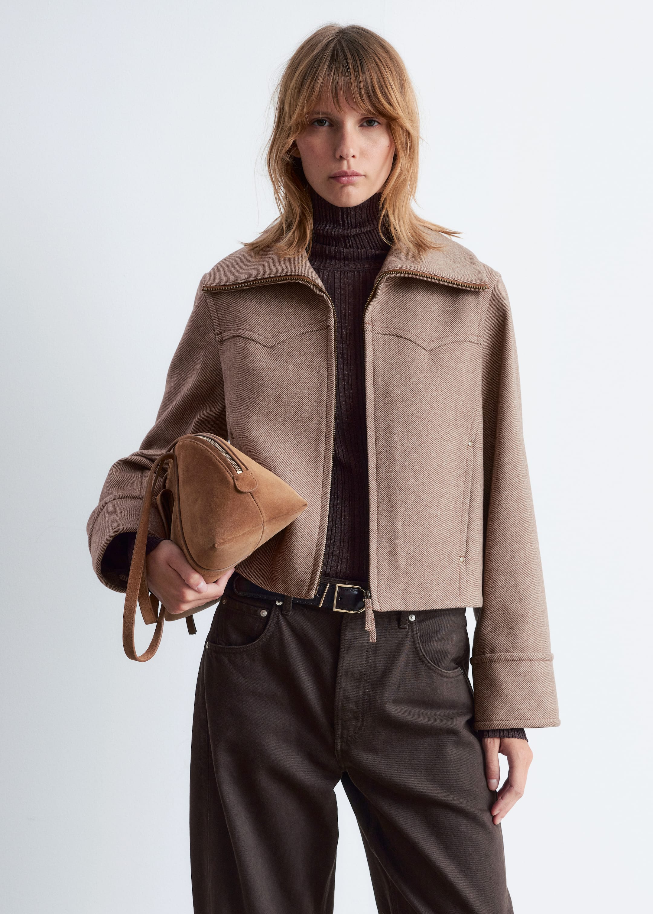 Größeres Bild ansehen: Eine hellbraune, kurze Jacke wird präsentiert, die ein strukturiertes Gewebe und einen Umlegekragen mit einem sichtbaren Frontreißverschluss aufweist, der bis zu diesem verläuft. Auffällige spitze Joche sind auf der Brust der Jacke sichtbar, die mit weiten, langen Ärmeln gestaltet ist und über einem dunkelbraunen gerippten Rollkragenpullover getragen wird.