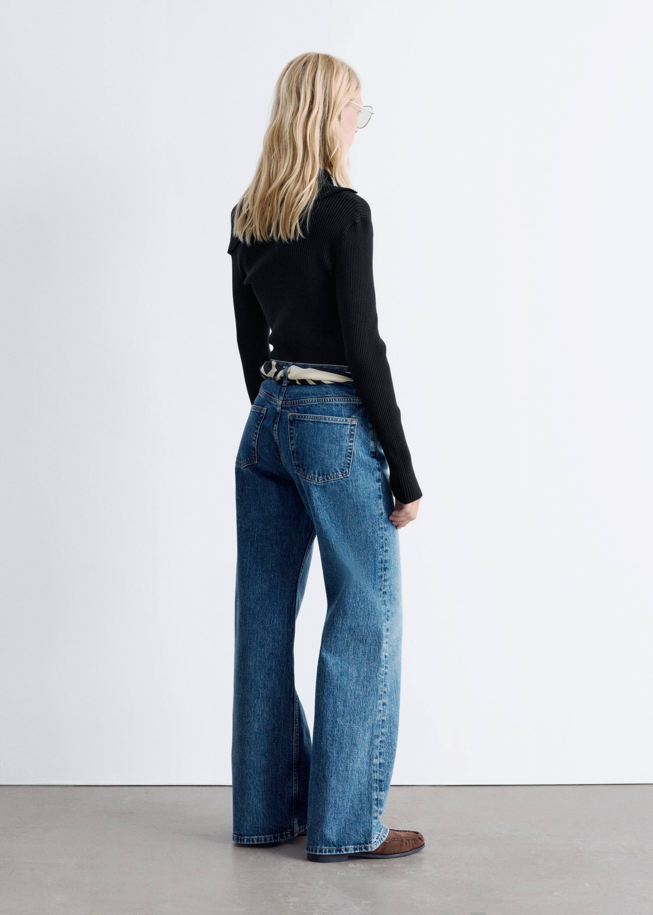 Grotere afbeelding bekijken: Een zwarte, lange mouw, geribbelde gebreide top met een slank, nauwsluitend silhouet wordt gedragen door het model. Een kraagstructuur is zichtbaar aan de achterkant van de nek, en de top is gestyled met blauwe wijde jeans.