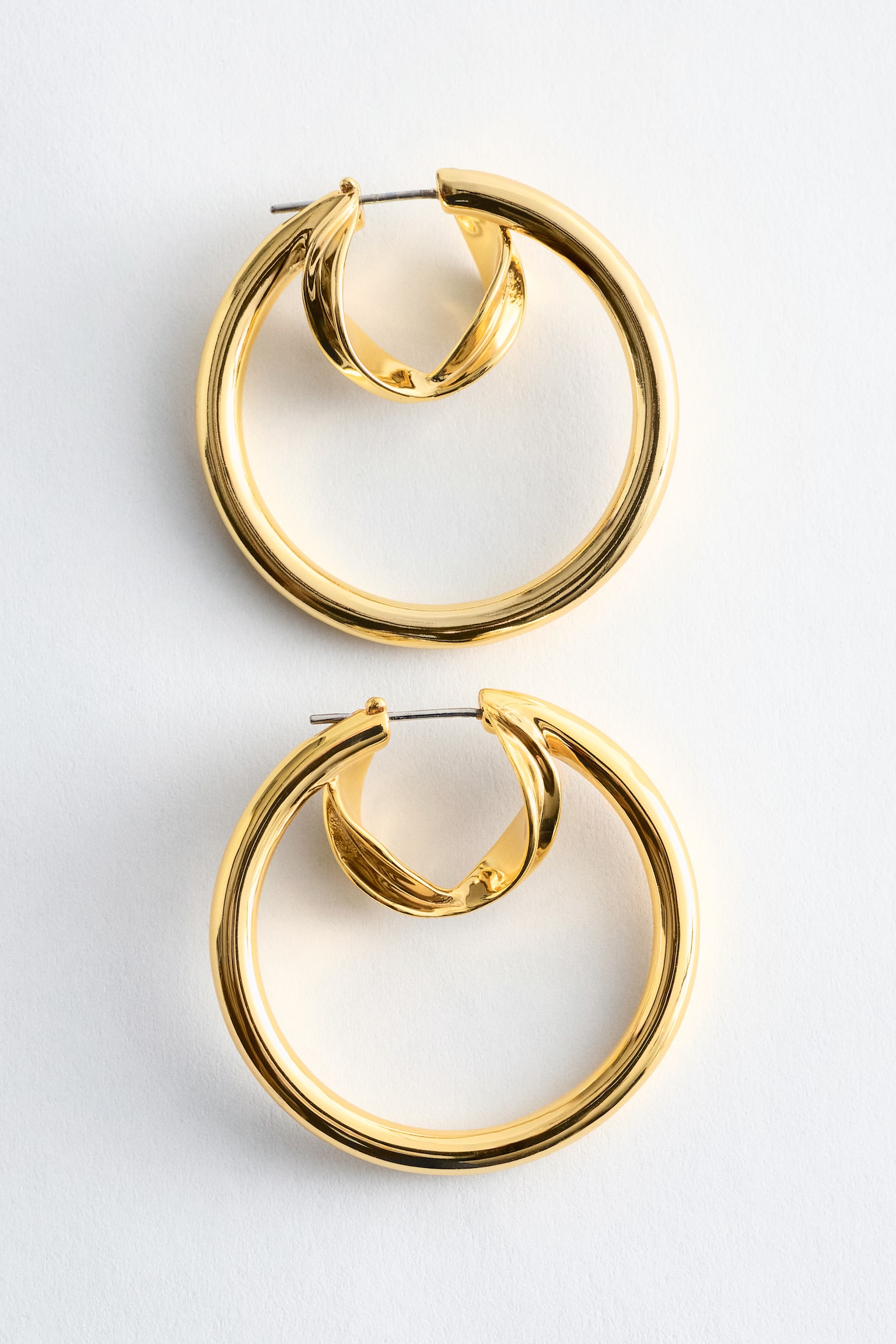Dois brincos de argola em latão polido são mostrados de frente, cada um apresentando uma grande argola exterior elegante que envolve uma argola menor embutida, definida por uma forma curva e escultural, com um fecho de encaixe visível no topo.