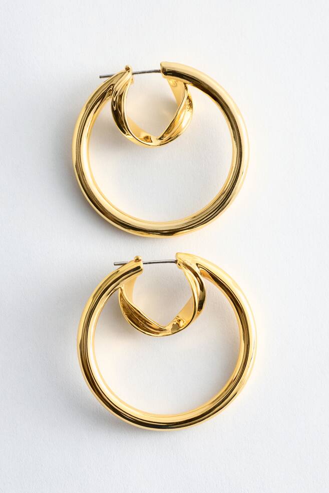 Two polished brass hoop earrings are displayed from the front, each showcasing a large, sleek outer hoop encasing a smaller, integrated hoop characterised by a curved, sculptural form, with a latch-back fastening visible at the top. - 1