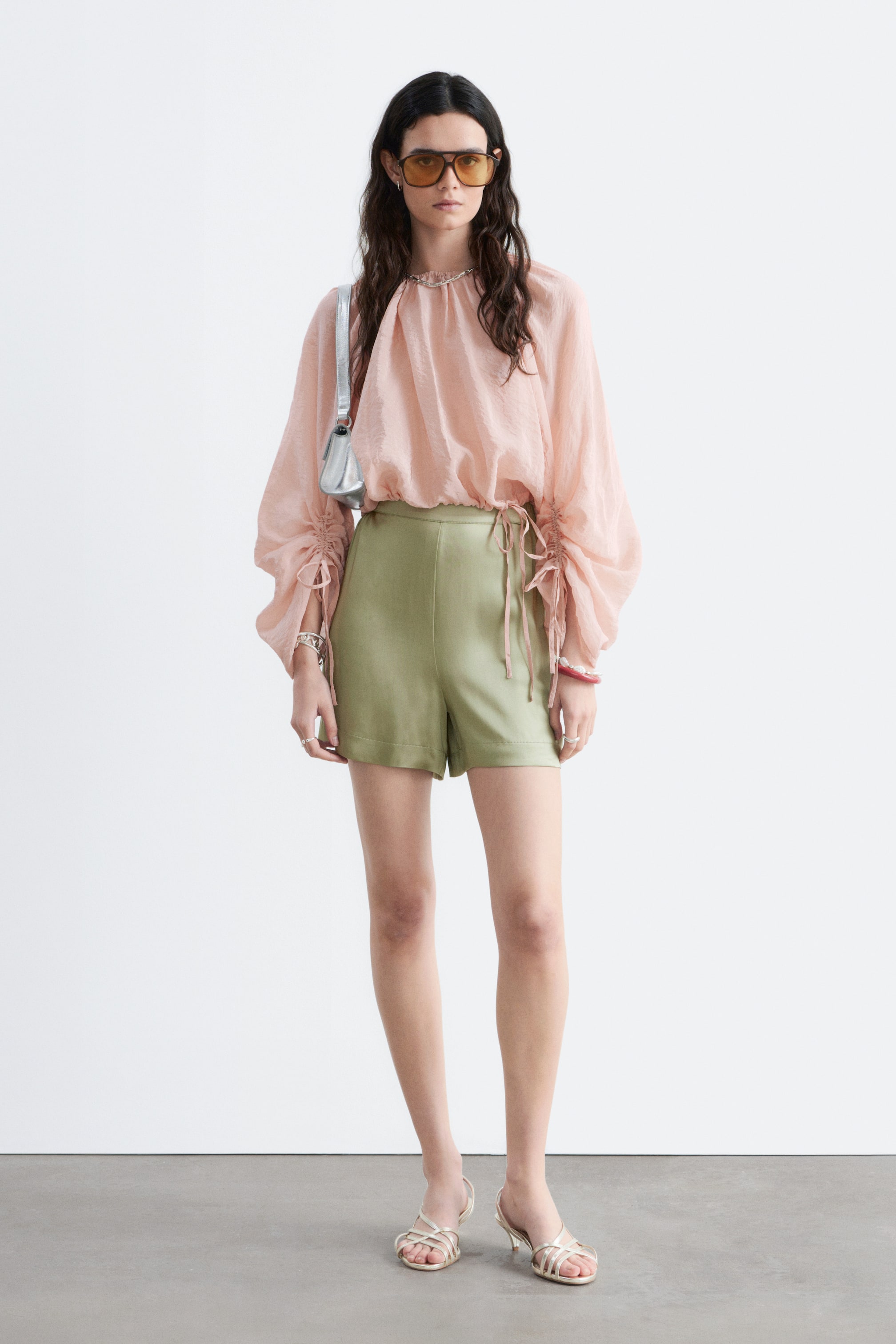 Agrandir l'image: La blouse rose pâle froissée présente un col montant à volants et des manches longues et volumineuses avec des poignets froncés et des liens, et son ourlet cintré est rentré dans un short vert olive.