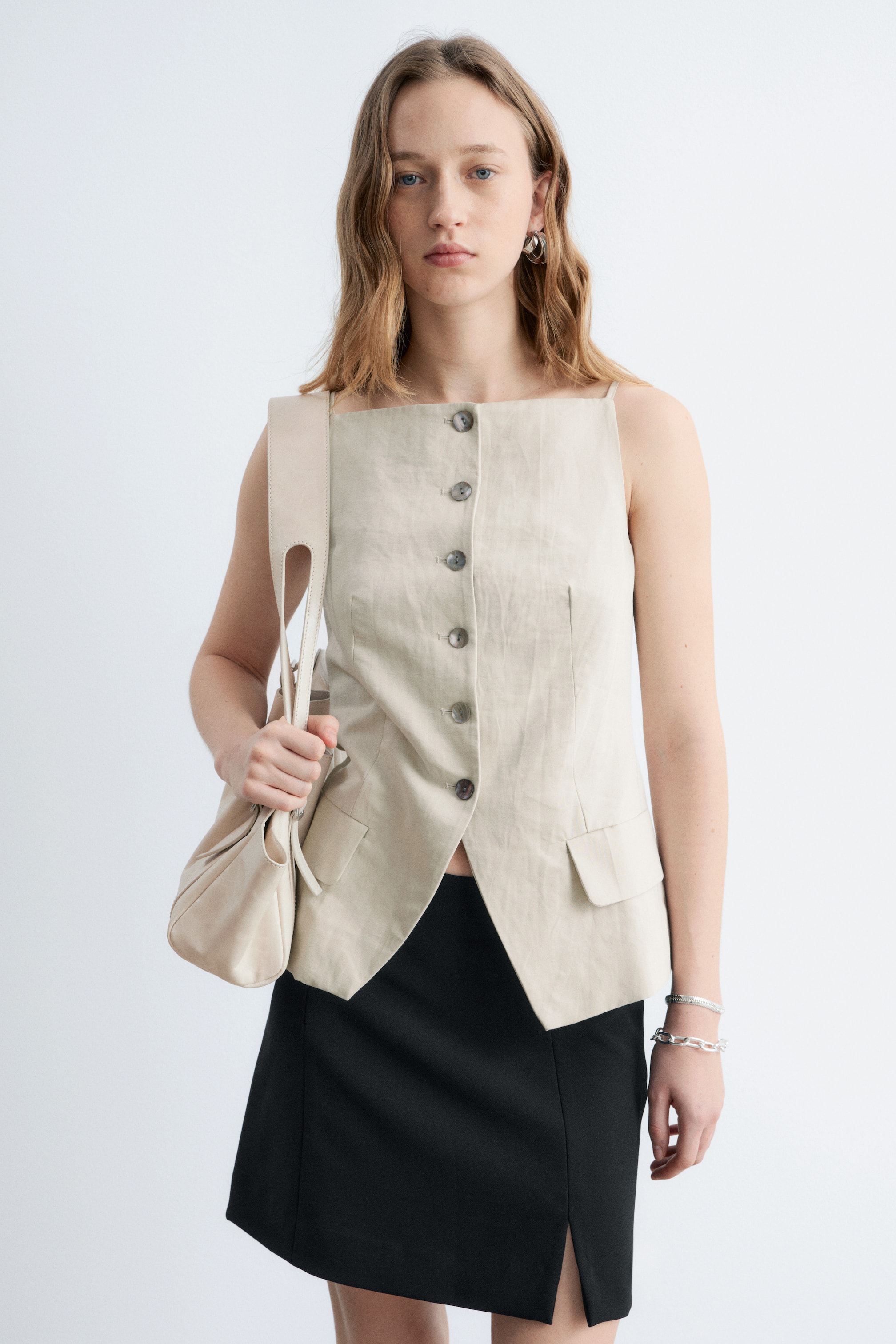 Größeres Bild anzeigen: Ein beigefarbener ärmelloser Weste mit quadratischem Ausschnitt, einer durchgehenden Knopfleiste mit mehreren dunklen Knöpfen und zwei Klappentaschen in der Nähe des Saums. Sie ist mit einem asymmetrischen V-förmigen Saum gestaltet.